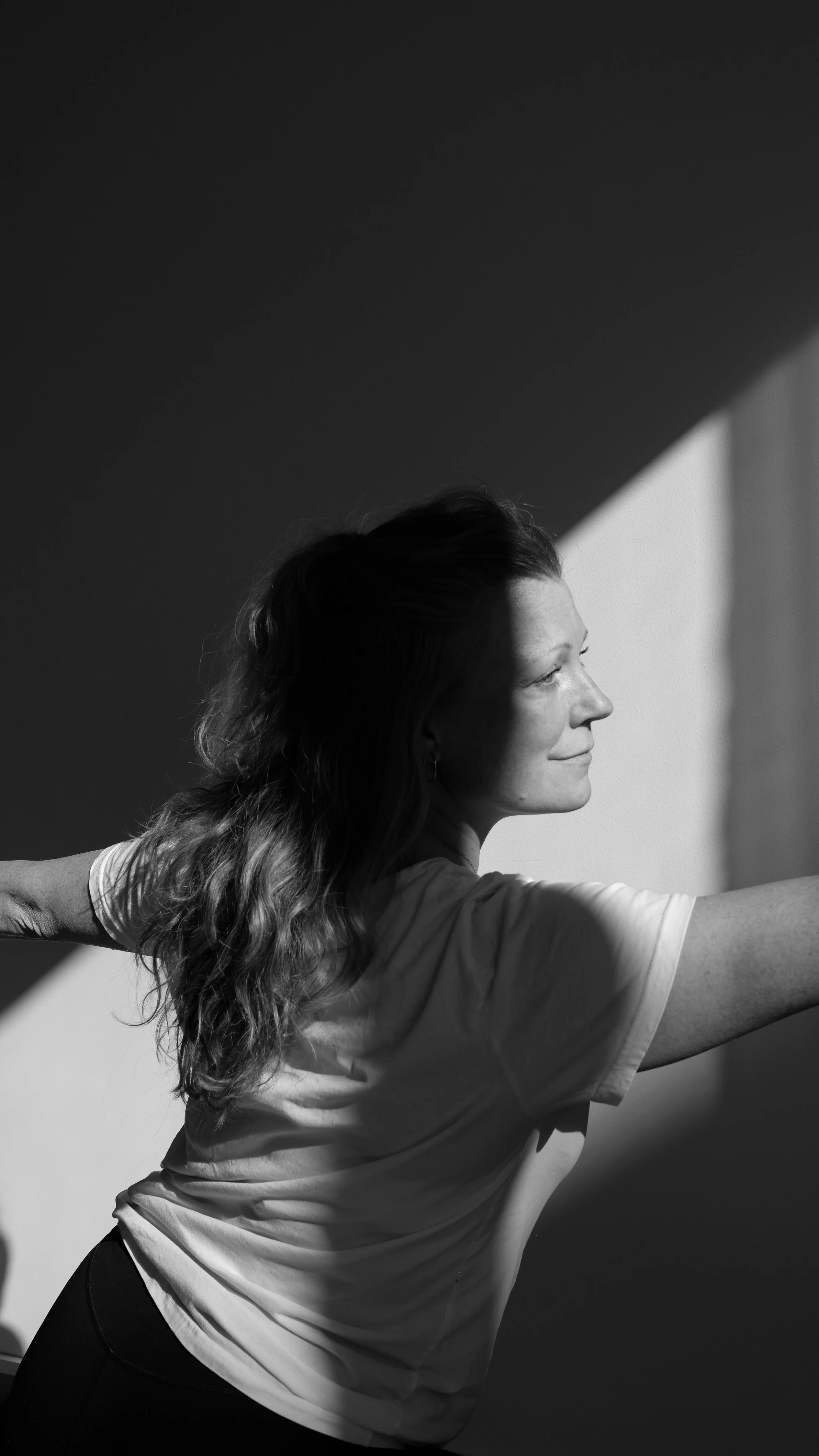 A woman with long hair stretching her arms to the sides in a yoga pose, standing beside a wall with sunlight creating angular shadows in a black and white photo.