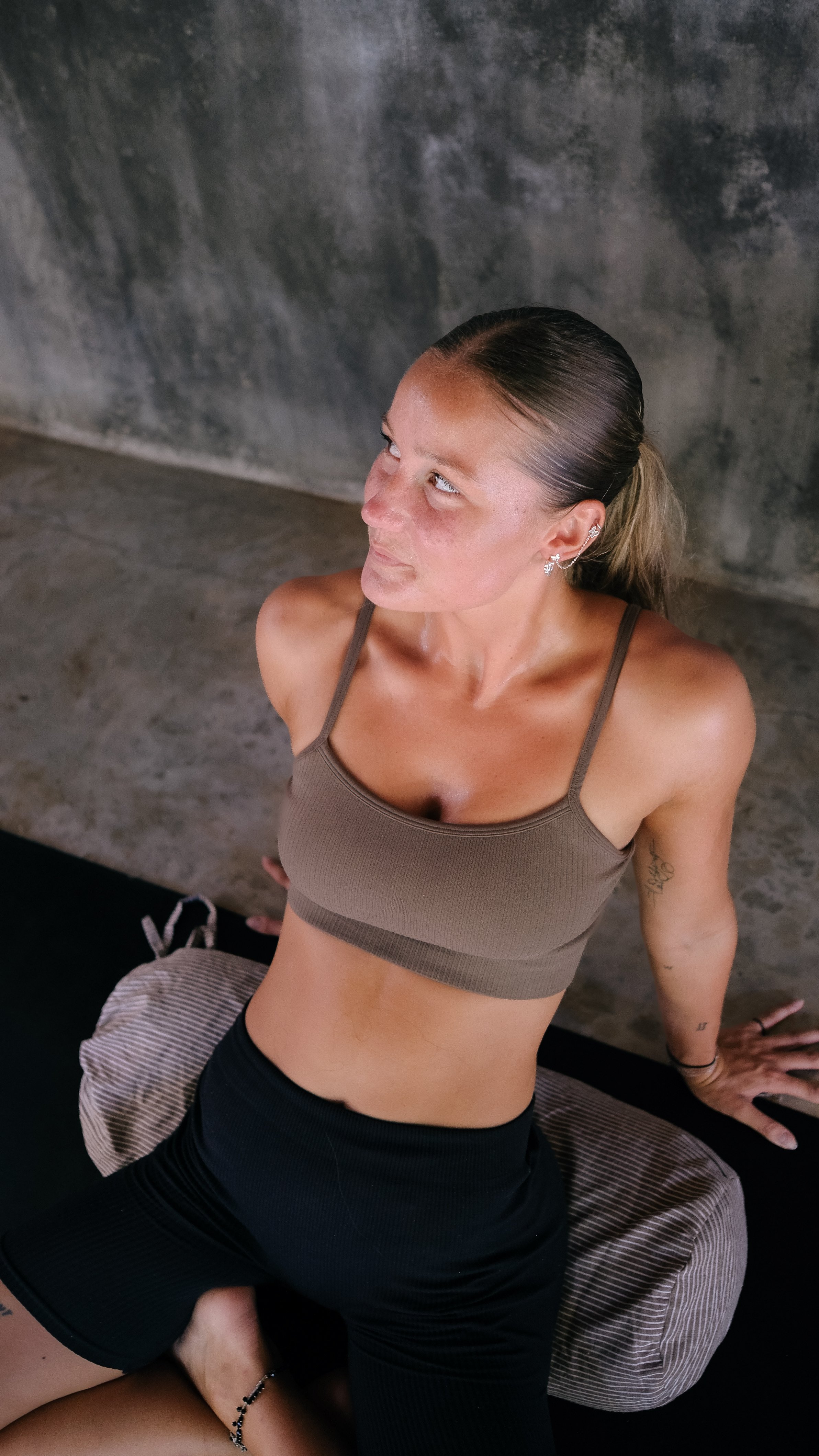 A woman with light skin, dark hair tied back, wearing a brown sports bra and black shorts, sitting on the floor with her back against a concrete wall, looking upward to her left with a relaxed expression.
