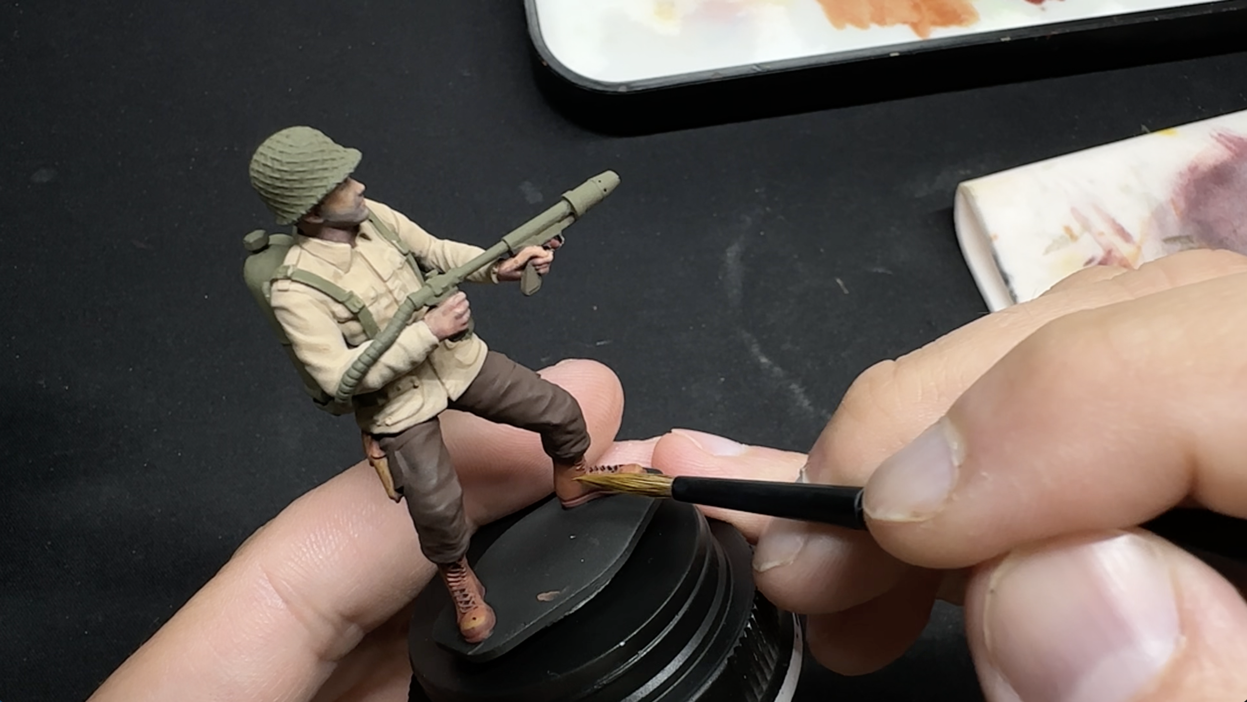A hand holding a paintbrush painting a miniature toy soldier dressed in military uniform, helmet, and carrying a rocket launcher against a black background.