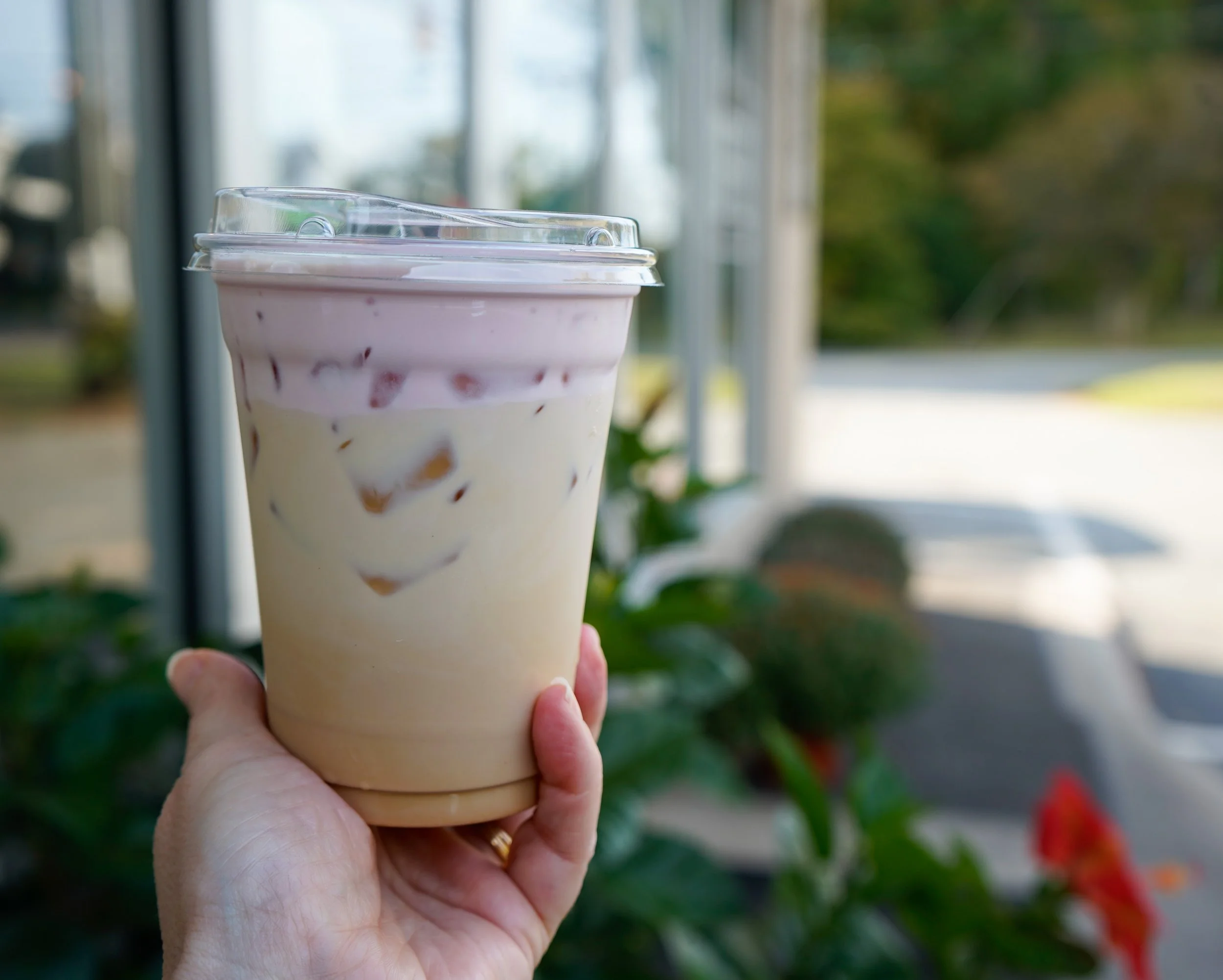 A hand holding a plastic cup filled with iced coffee or frappuccino with cream and ice, inside a cafe or shop with large windows and some outdoor plants and flowers.