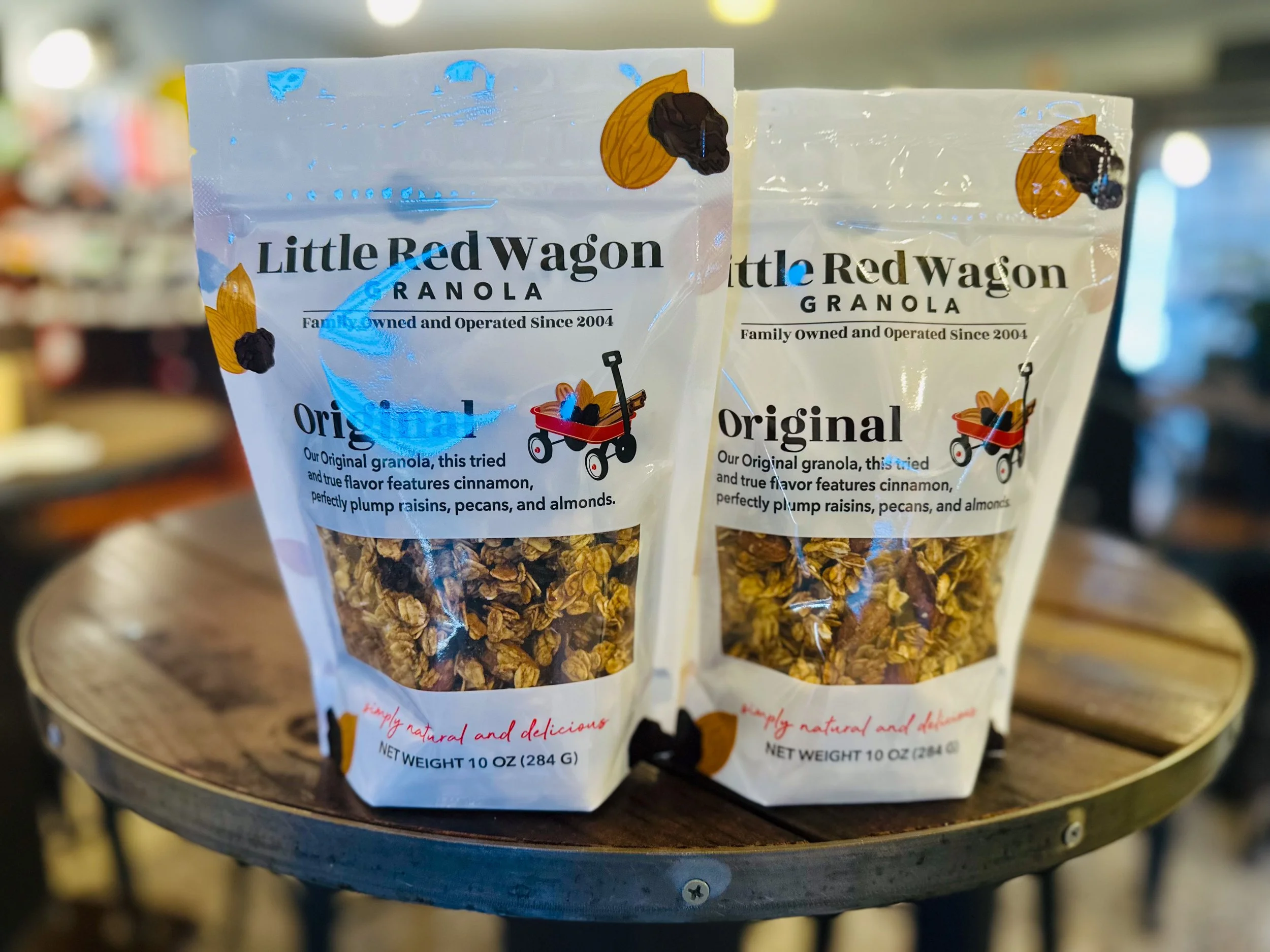Two bags of Little Red Wagon Granola on a wooden table in a grocery store, with a blurred background.
