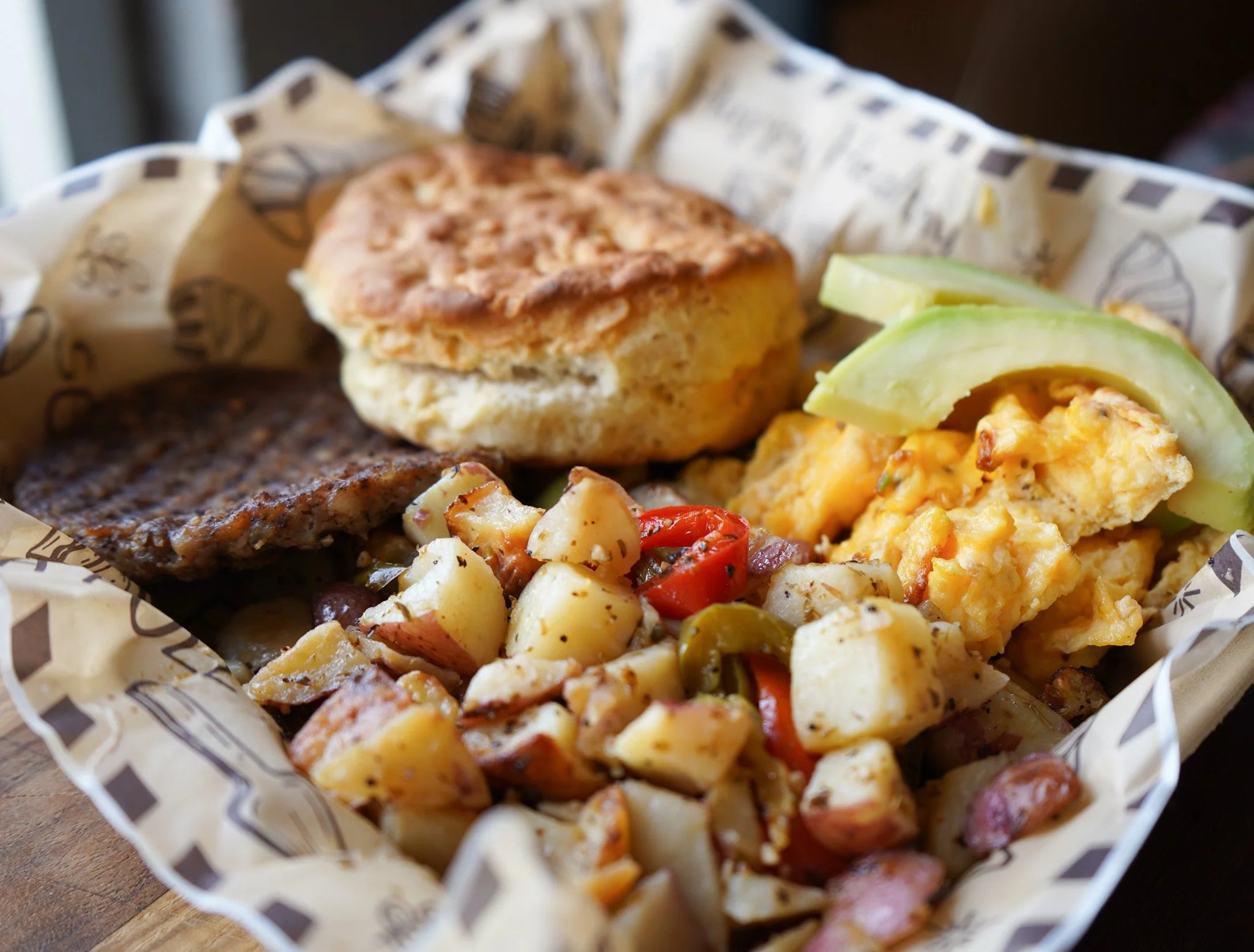 A basket of breakfast food includes scrambled eggs topped with avocado slices, a sausage patty, a biscuit, and roasted potatoes with red and green peppers.