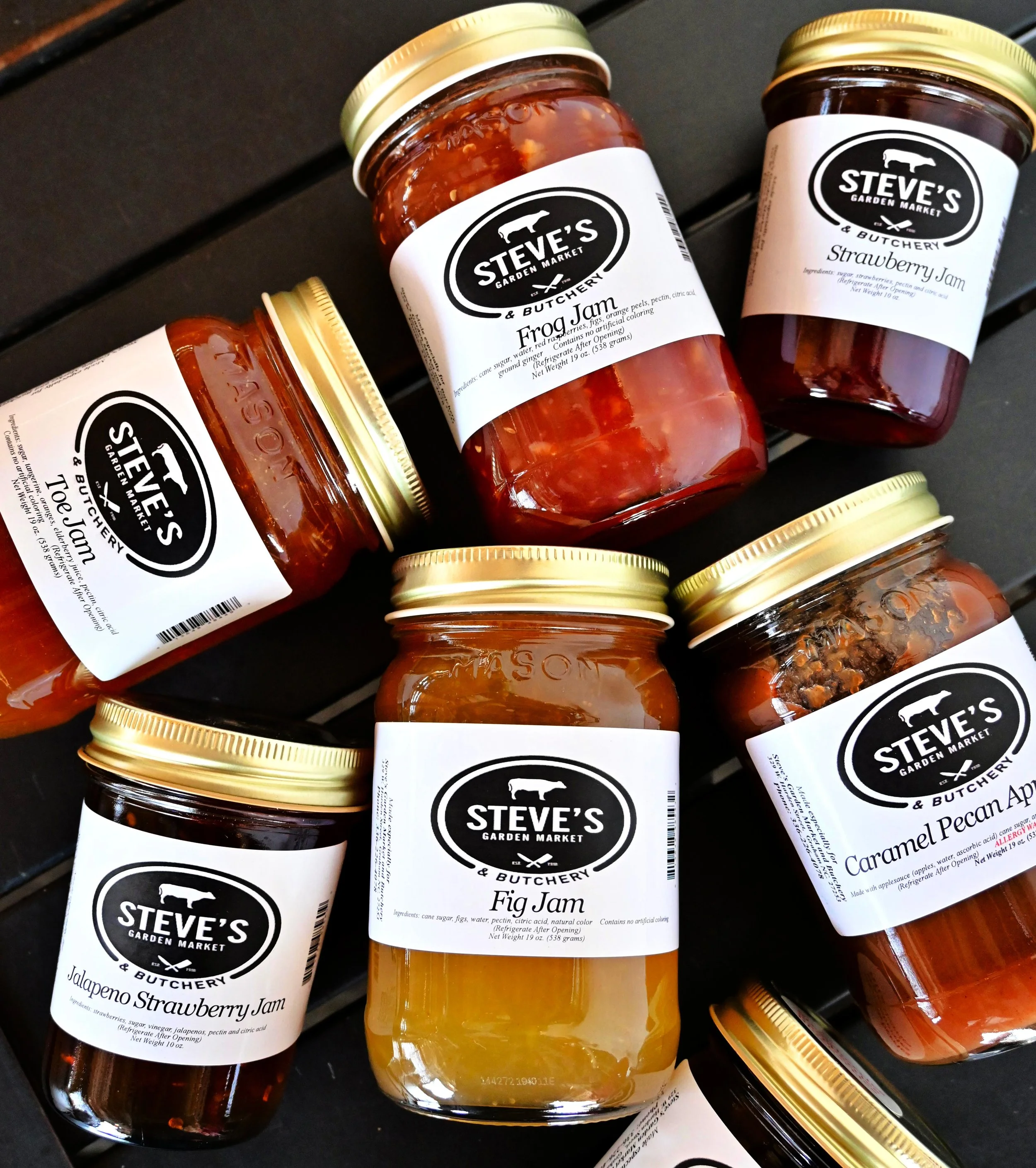 Assorted jars of jams and spreads from Steve's Garden Market & Butchery, including frog jam, strawberry jam, jalapeno strawberry jam, fig jam, caramel pecan apple jam, and strawberry jam, on a black surface.