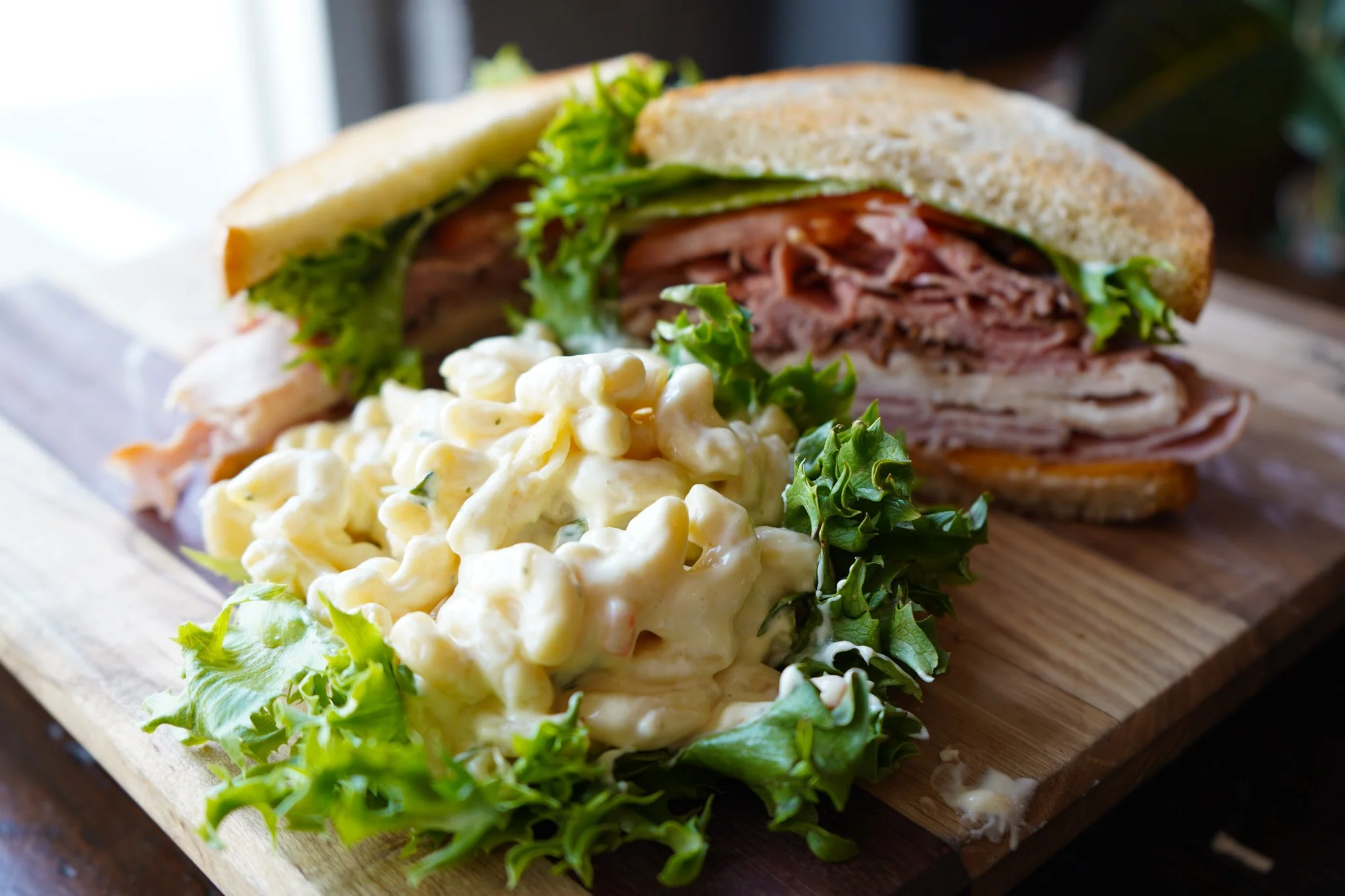 A sandwich with sliced meat and lettuce on whole grain bread, served with a side of macaroni salad on a wooden cutting board.