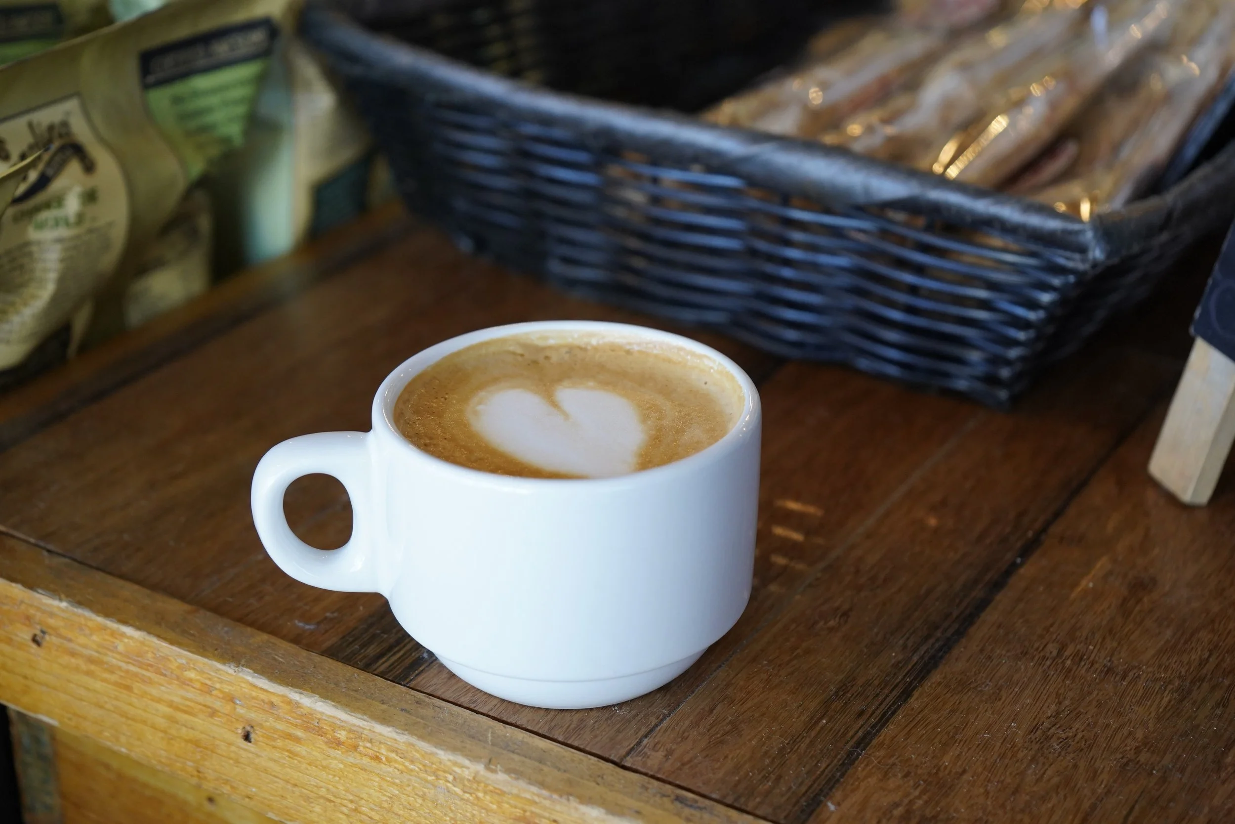A white ceramic cup of coffee with a heart-shaped latte art on top, placed on a wooden table with a black basket and packaged snacks in the background.