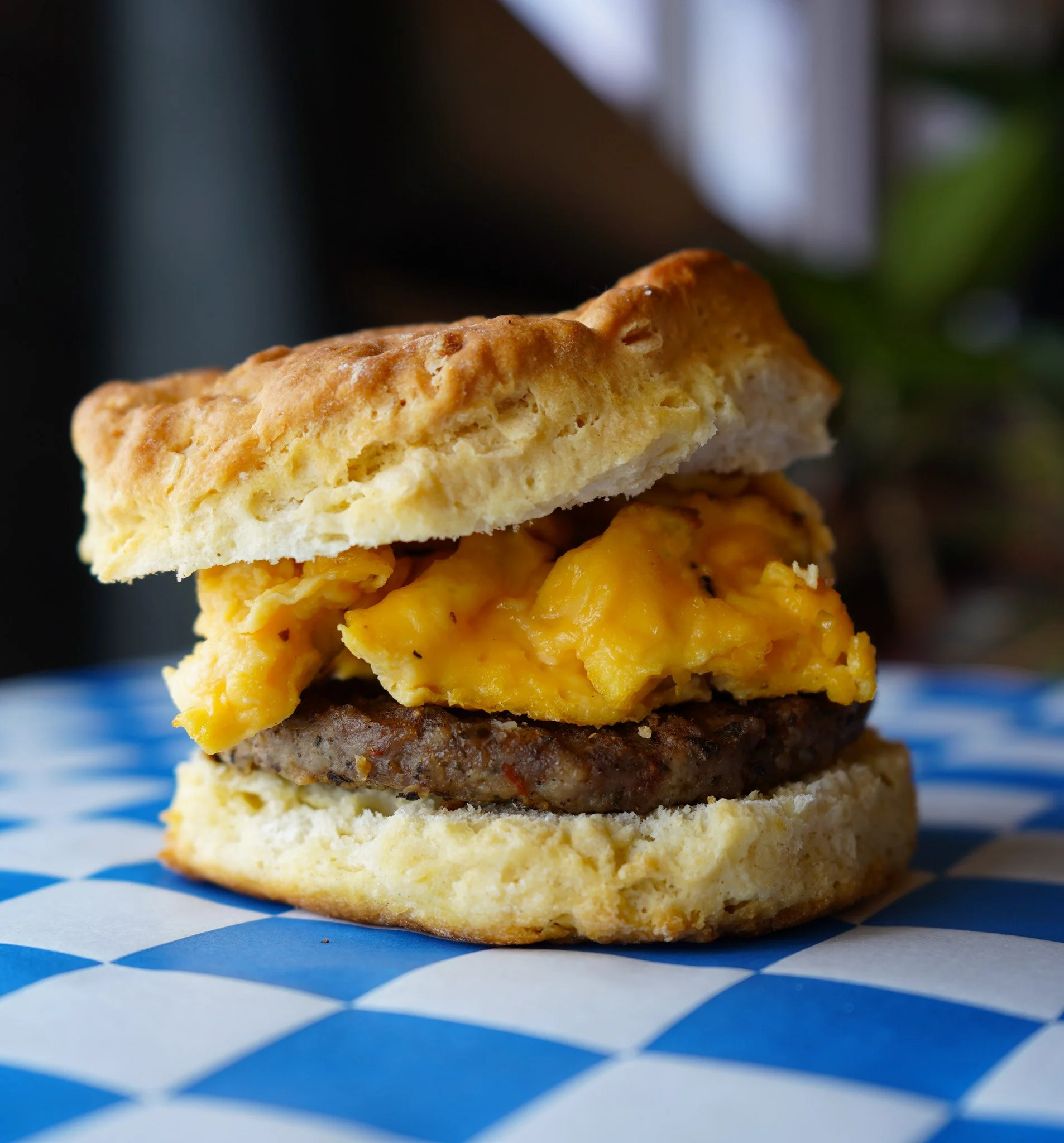 Close-up of a breakfast sandwich featuring scrambled eggs, a sausage patty, and a biscuit bun, placed on a blue and white checkered paper