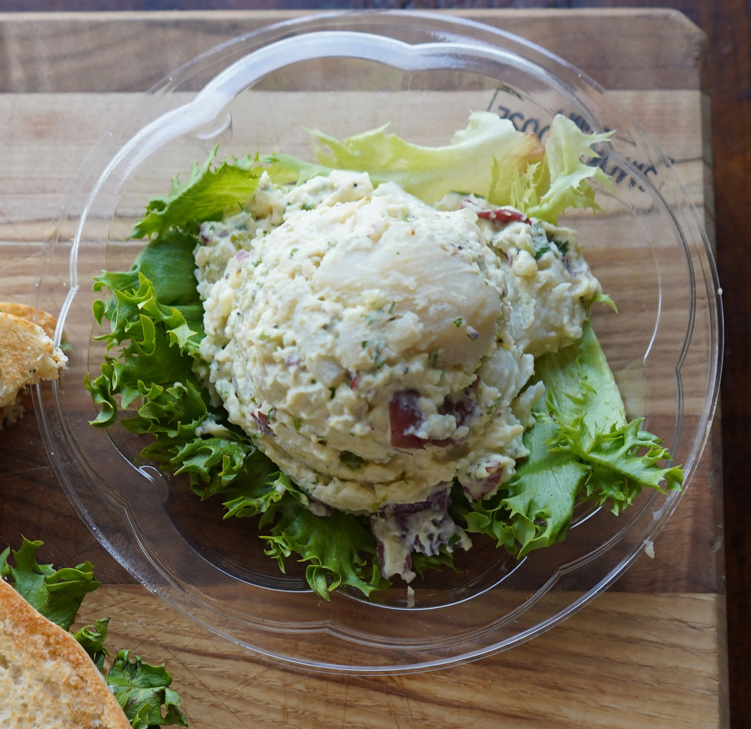 A serving of potato salad with chopped vegetables on lettuce leaves inside a clear plastic container, placed on a wooden surface.