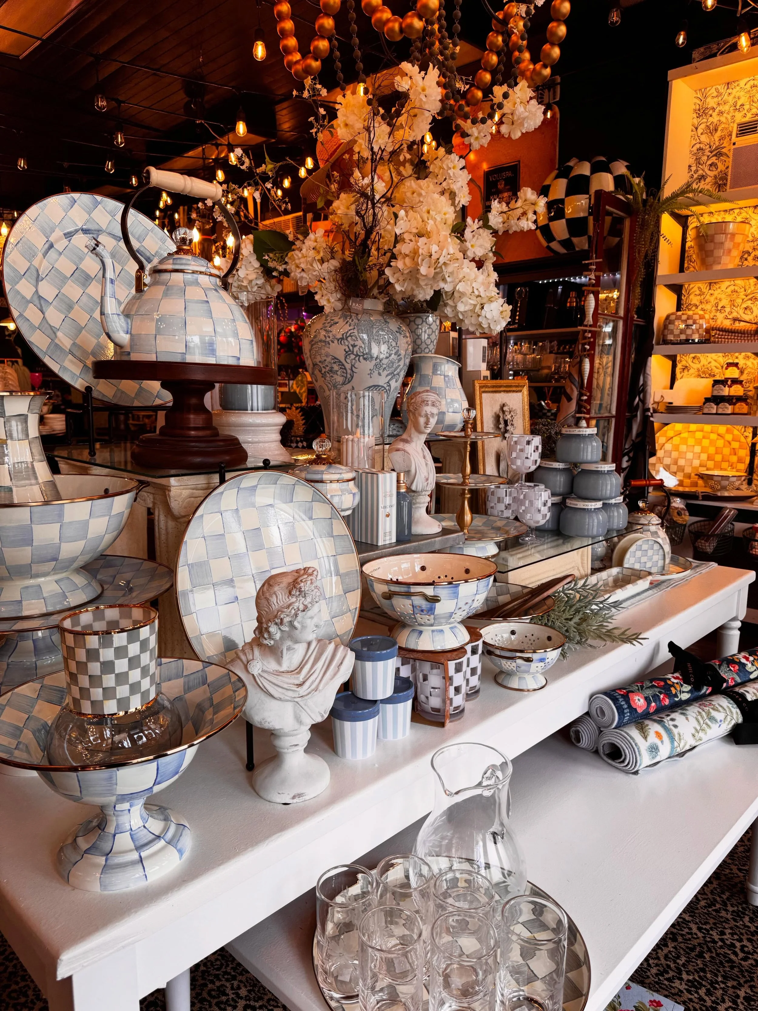 Display of patterned dishware, vases, and decorative items on a white table in a home decor store, with floral arrangements and decorative objects.