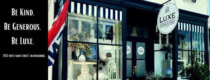 Storefront of Luxe Floral & Home, with black and white striped awnings and a sign, showcasing floral arrangements inside. A black sign on the left displays the words 'Be Kind. Be Generous. Be Luxe' and the address 302 West Main Street, Bloomsburg.