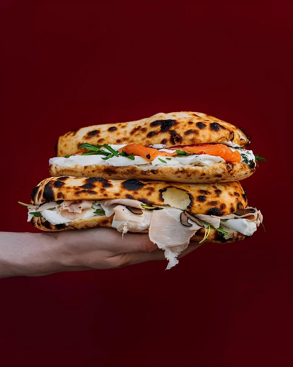 Un sandwich géant avec deux morceaux de pain grillé, garniture de saumon, de poulet, d'herbes, et de fromage, tenu dans une main contre un fond rouge.