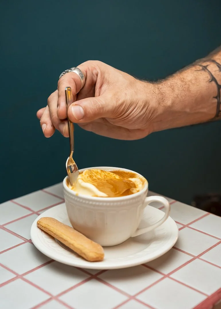 Une main tenant une cuillère au-dessus d'une tasse de café crème avec une biscotte sur une assiette sur une table à carreaux
