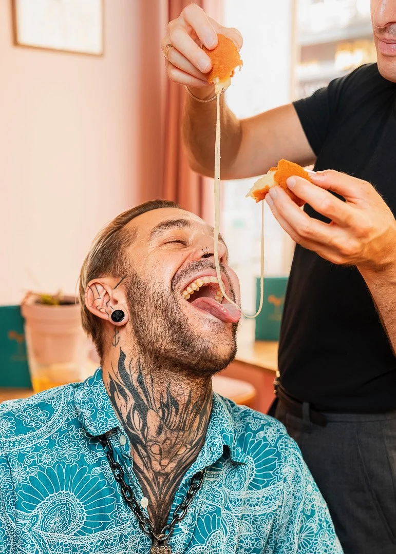 Un homme avec des tatouages et des piercings ouvre grand la bouche pendant qu'une autre personne lui verse de la sauce sur la langue avec un morceau de pain.