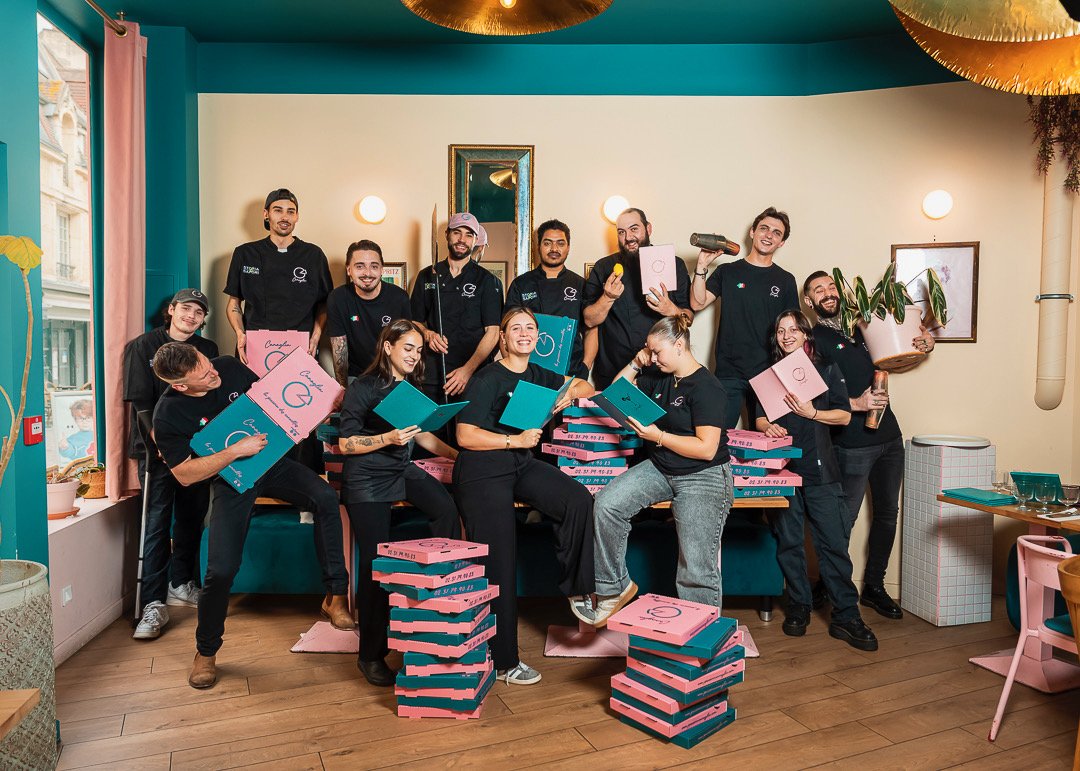 Groupe de personnes souriantes dans un intérieur coloré, entouré de boîtes à pizza empilées, tenant des livres ou documents et posant pour une photo de groupe.