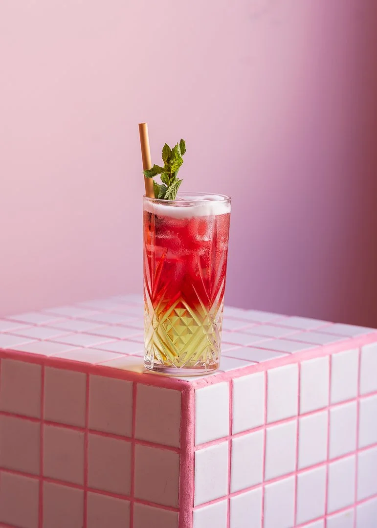 Un cocktail coloré dans un verre décoratif avec une feuille de menthe et une paille en bambou, placé sur une table en carreaux roses et blancs.
