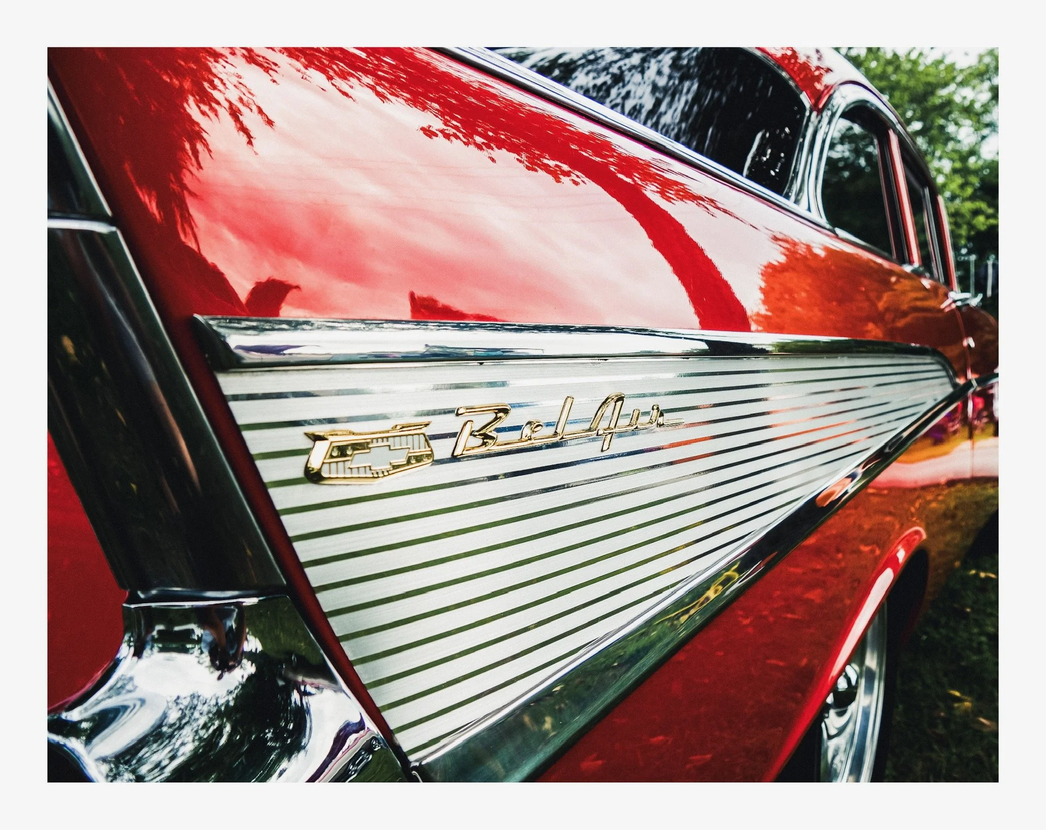 Close-up of a vintage red Chevrolet Bel Air, showcasing the front grille with chrome details and the Chevrolet emblem, parked outdoors with trees in the background.