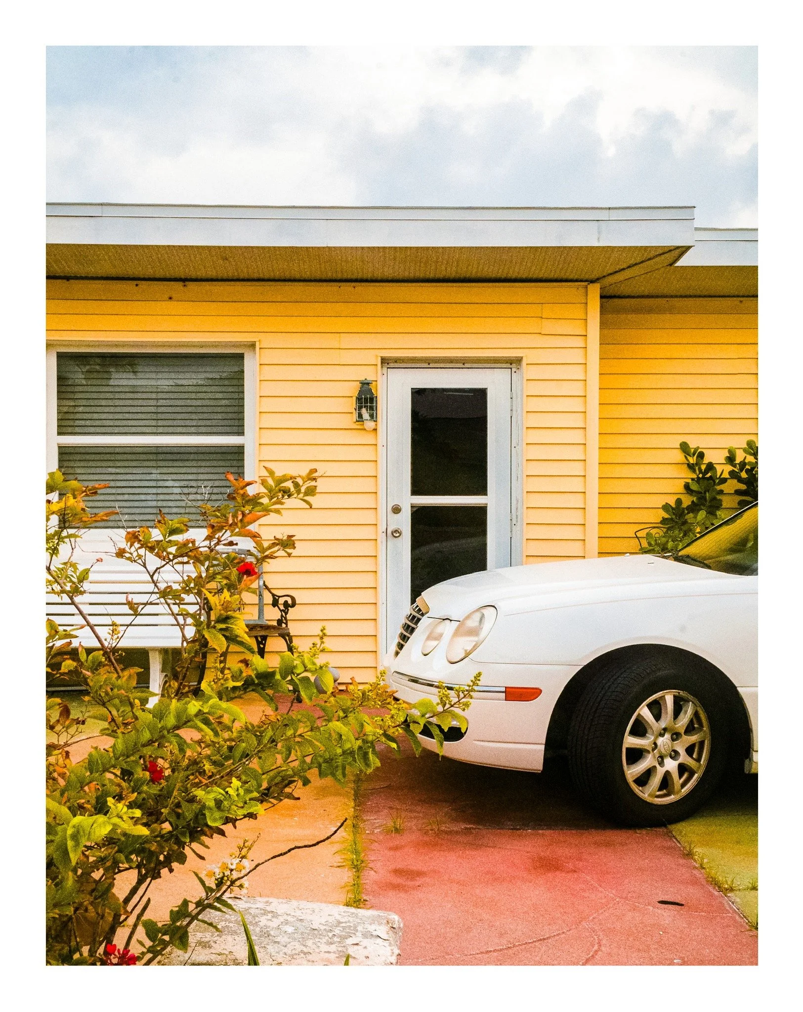 Part of a white car parked in front of a yellow house with a white door, a window with blinds, and a wall-mounted lantern, with plants and bushes in the foreground.