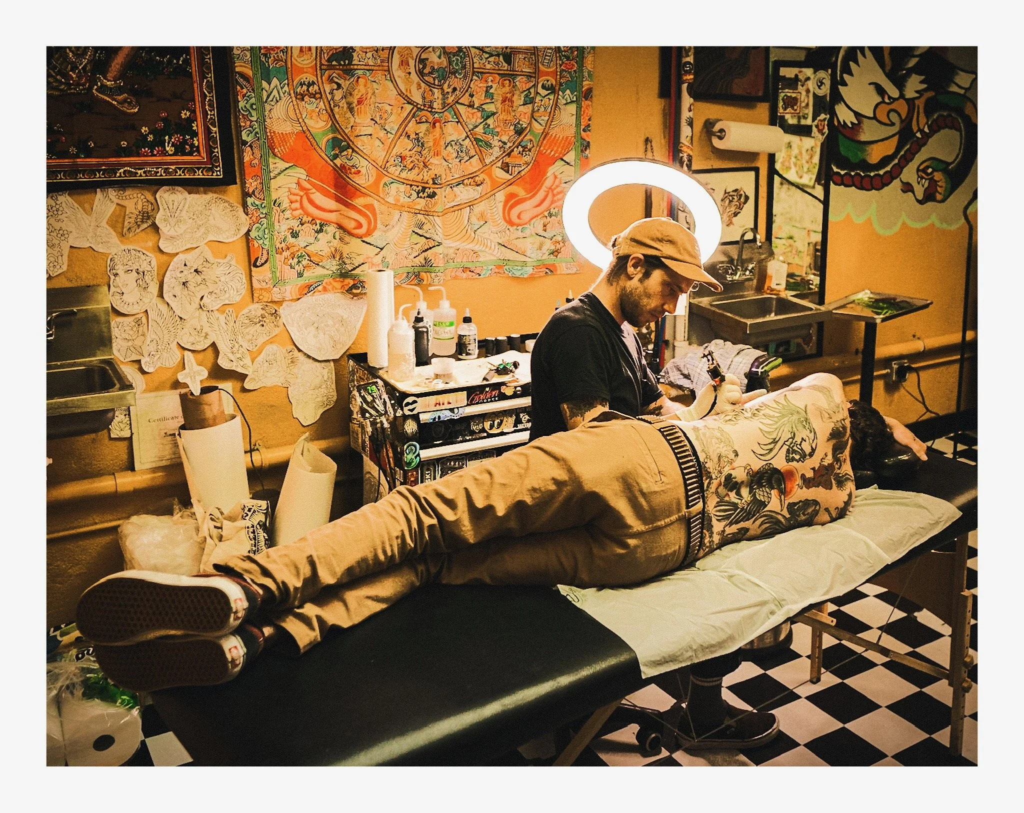 A tattoo artist working on a client's leg in a tattoo studio decorated with artwork and hanging tapestries, with tattoo supplies on a table and various drawings on the wall.