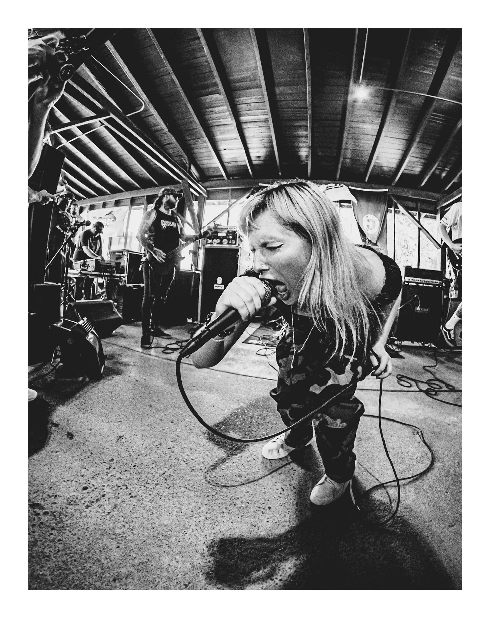 A black and white photo of a woman singing into a microphone, wearing camouflage pants and sneakers. Behind her, a band plays on a stage with wooden ceilings and large windows, possibly in a cozy indoor venue.