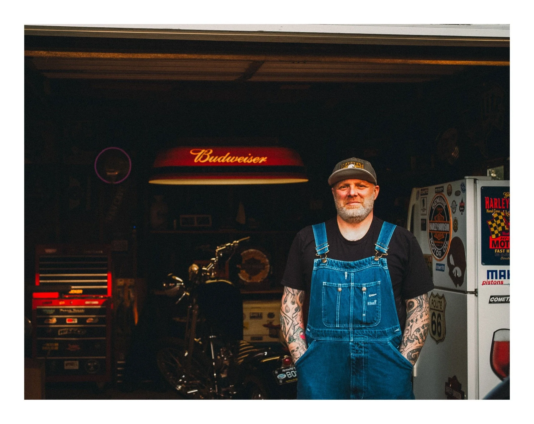 Man with tattoos on arms, wearing a gray cap and blue overalls, standing inside a garage with motorcycle parts and signage, including a Budweiser sign above his head.