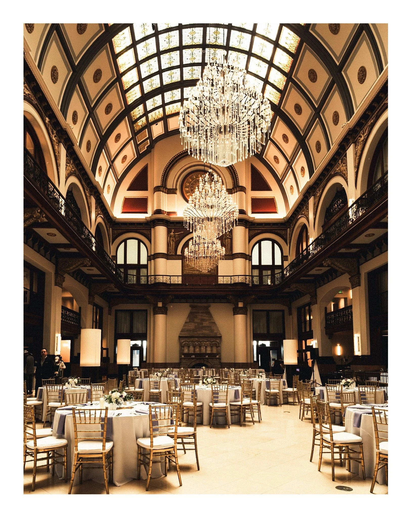 Elegant banquet hall decorated for a formal event with round tables, white tablecloths, floral centerpieces, and gold Chiavari chairs. The high ceiling features large chandeliers and stained glass windows, with an ornate balcony rail around the upper