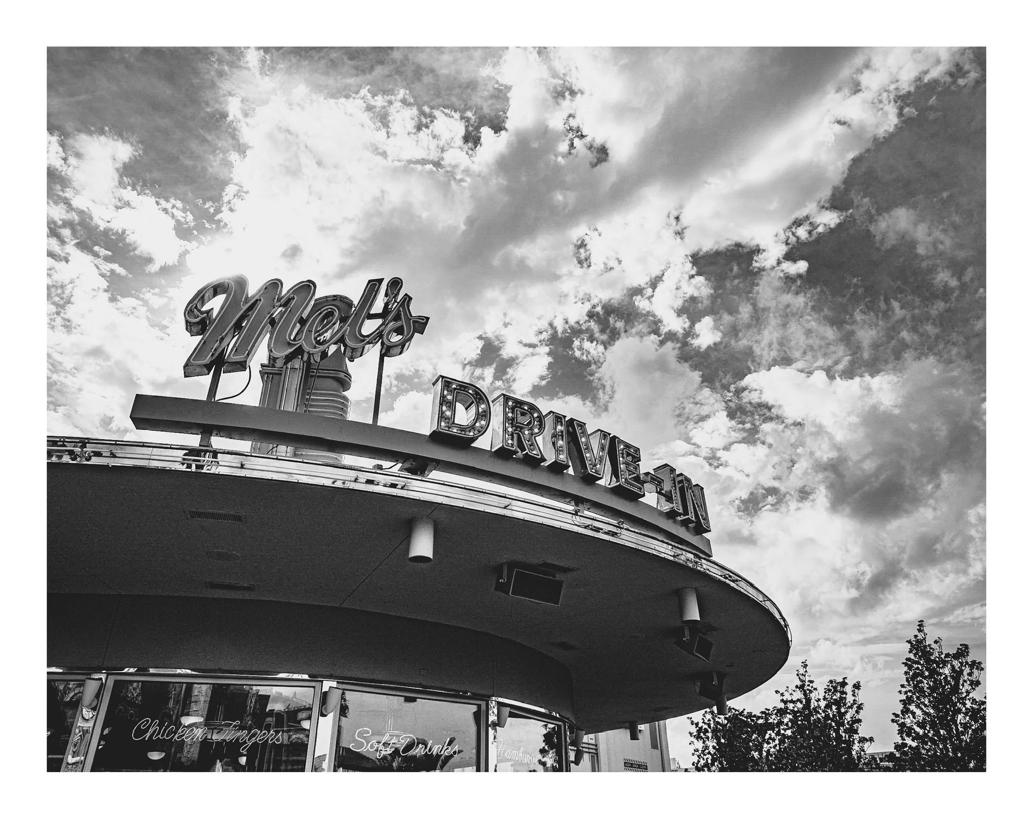A black and white photograph of a large sign that says "Mots" with a sky filled with clouds in the background. Part of a building with neon signs advertising chicken tenders, soft drinks, and other foods is visible below the sign.