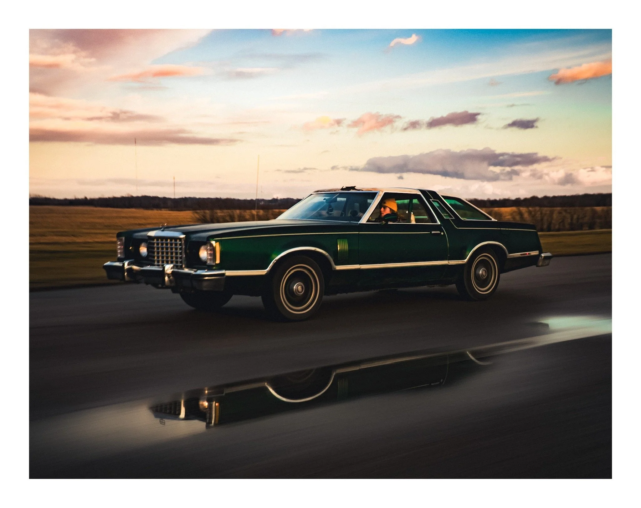 A woman wearing a yellow hat driving a vintage dark green car on an open road during sunset, with a colorful sky and clouds.
