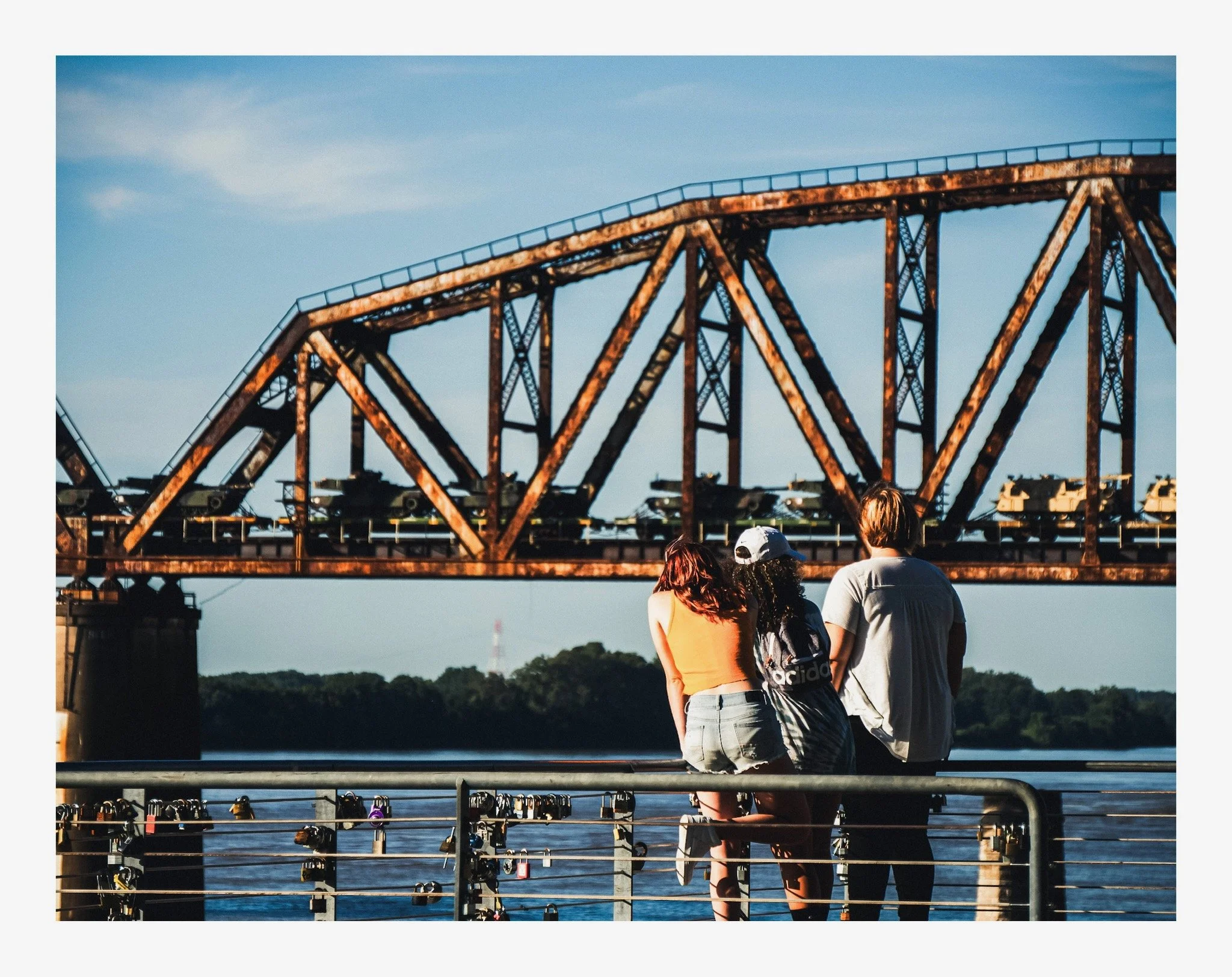 Several people standing near a waterfront, facing a rusted metal bridge structure with a blue sky above.