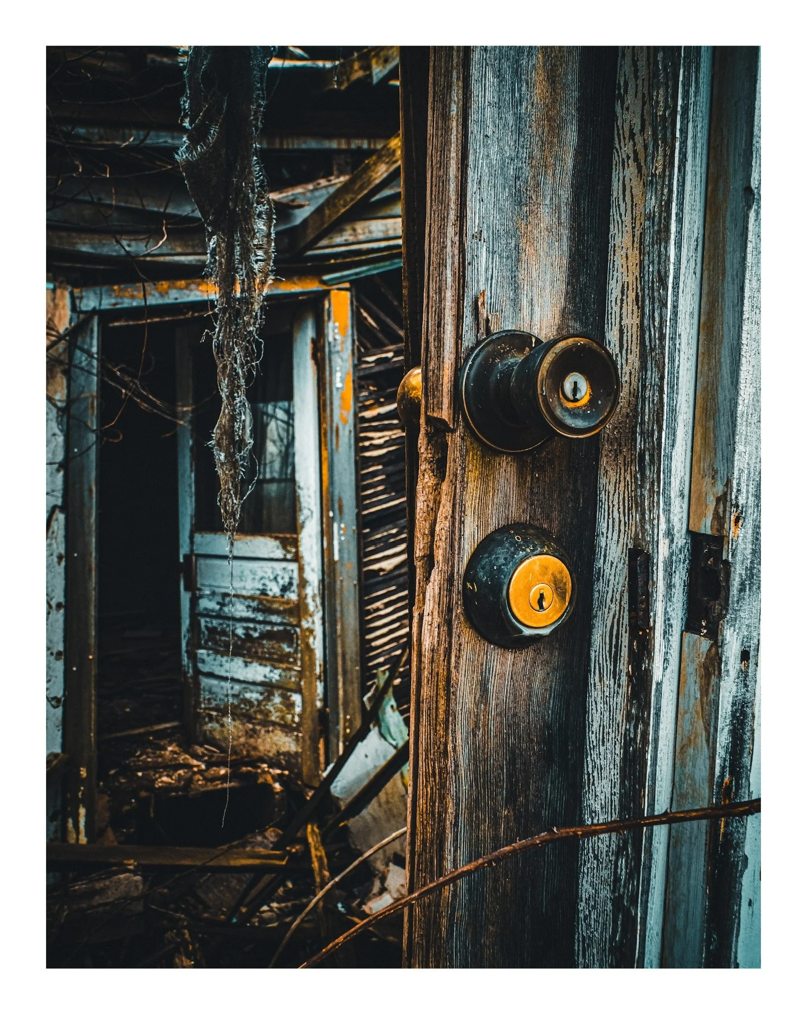 Close-up of a weathered wooden door with a brass doorknob and keyhole, part of a dilapidated structure with peeling paint and broken windows, surrounded by debris and tangled branches.
