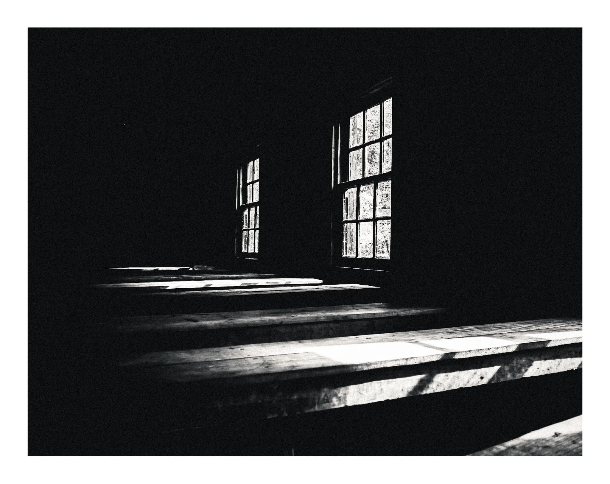 Black and white photograph of interior with large windows, wooden stairs, and shadows on the floor.