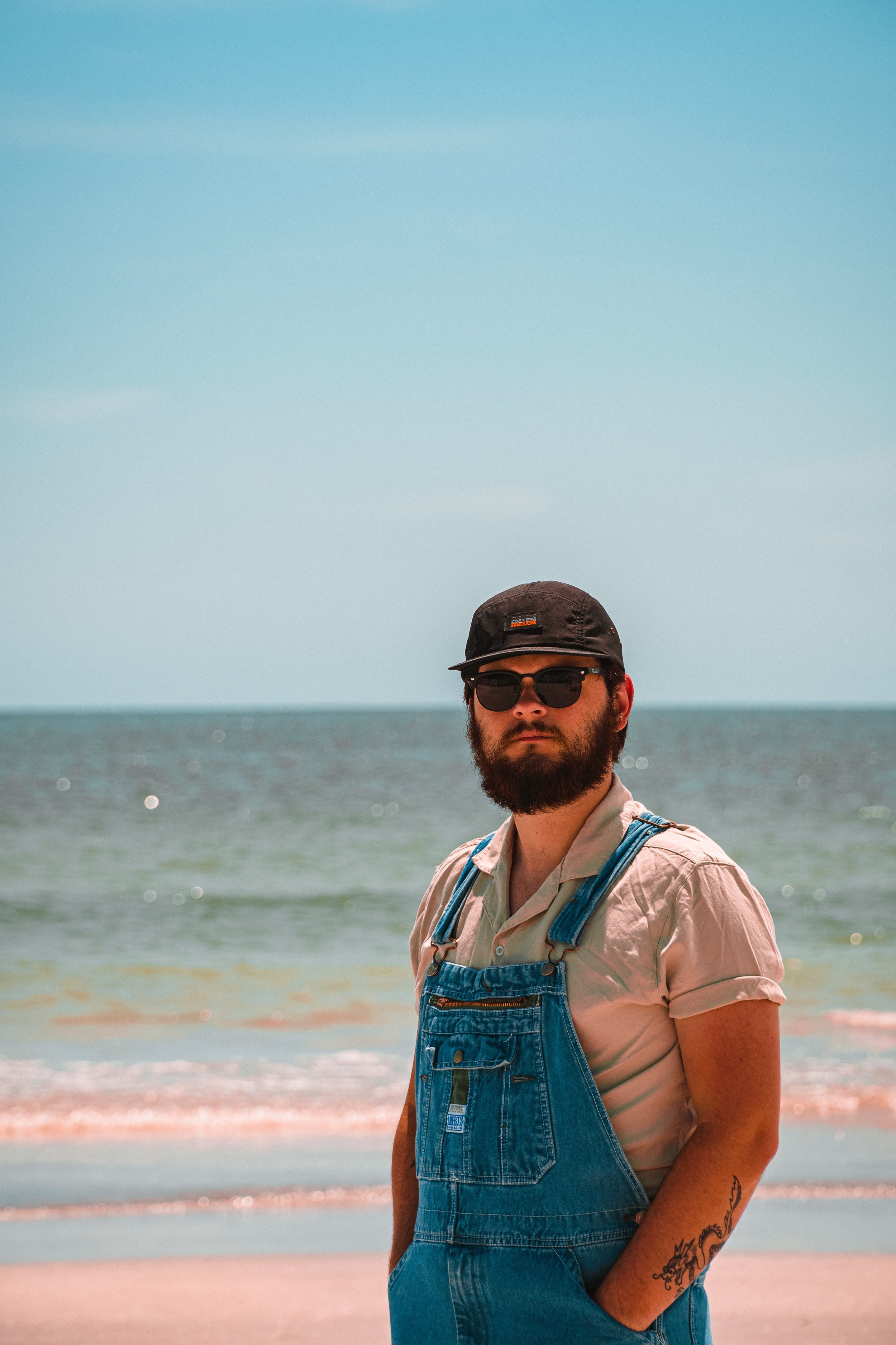 A man with a beard and tattoos on his arm standing on a beach with a blue sky and ocean in the background, wearing a cap, sunglasses, a beige shirt, and denim overalls.