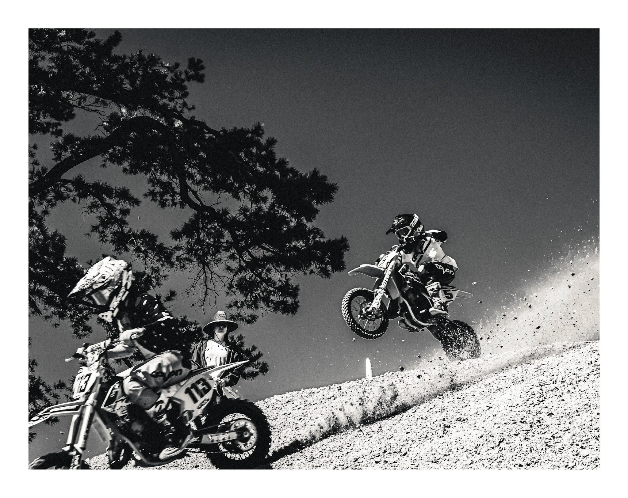 Black and white photo of mountain bikers riding on a dirt trail beneath a tall pine tree at night, with a clear starry sky.