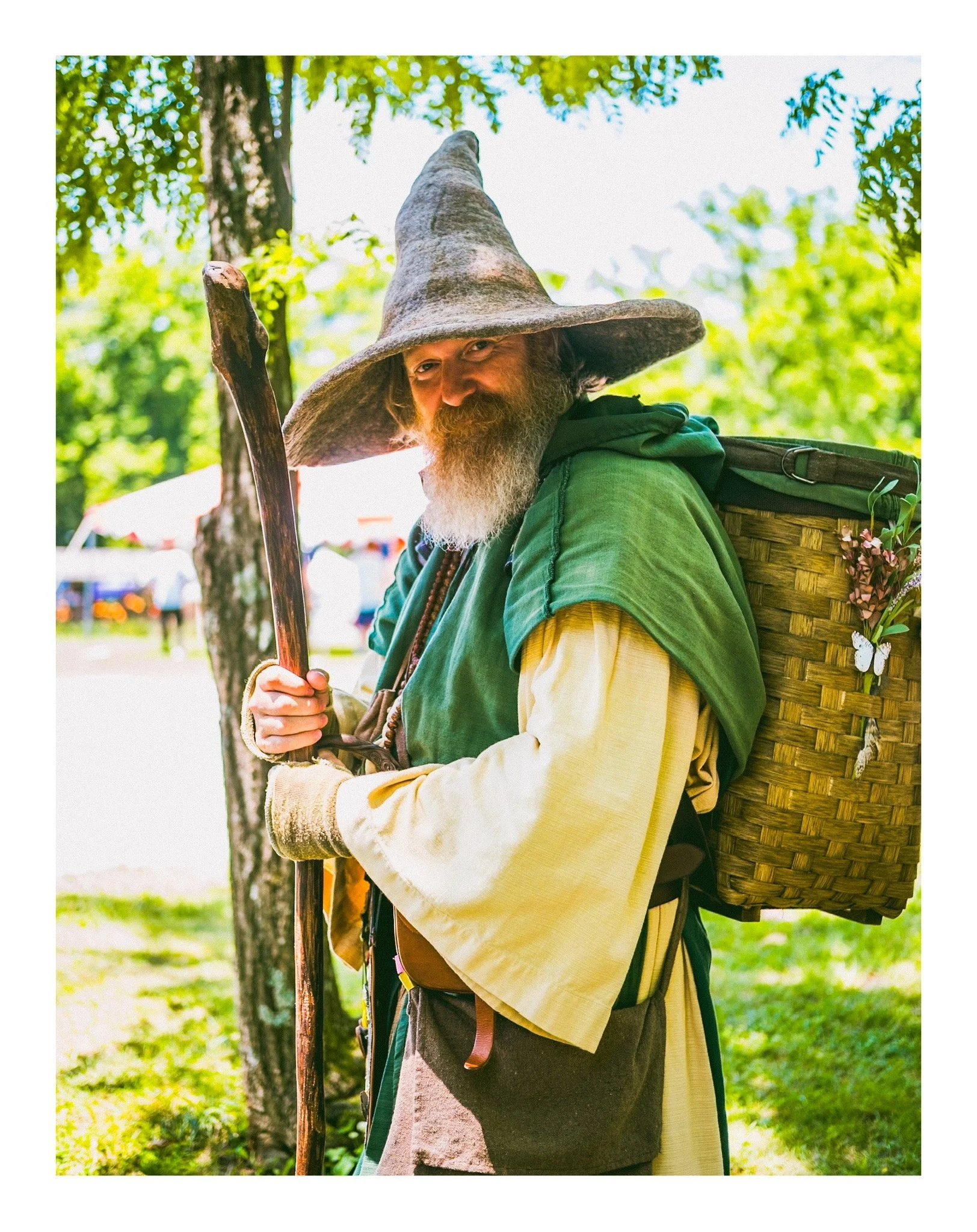 Man dressed as a wizard with a large, pointed hat made of rough material and a long beard, holding a wooden staff, outdoors with trees and bright sunlight in the background.