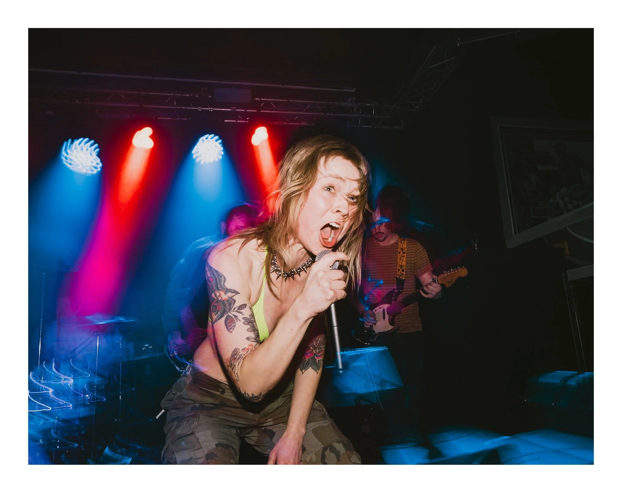 A woman singing passionately into a microphone during a live music performance. She has long hair, tattoos on her arms, and is wearing a sleeveless top and camouflage pants. The background features colorful stage lighting with blue and red lights.