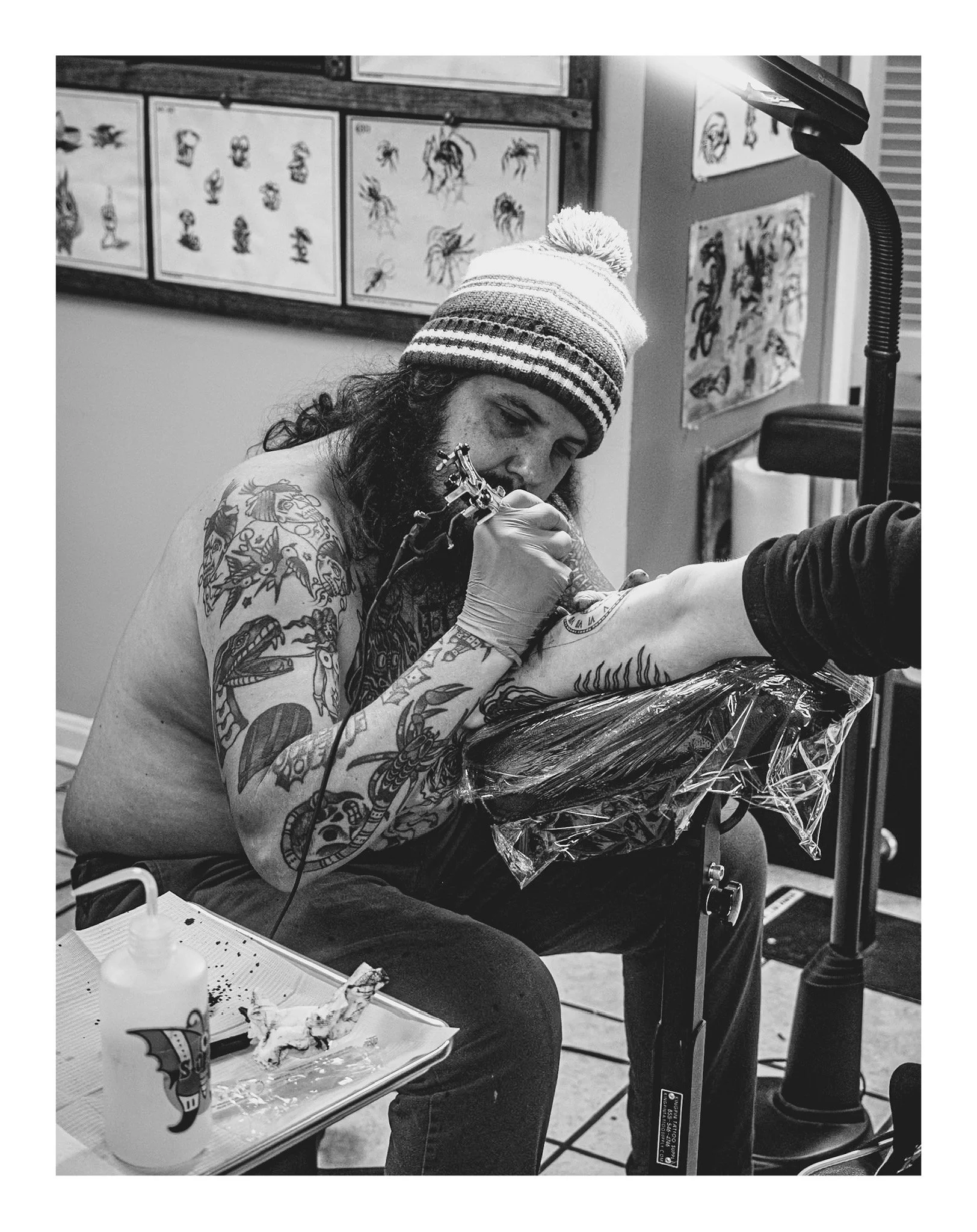 A person with tattoos on their arm, wearing a striped beanie, is getting a tattoo on their forearm at a tattoo studio. There are tattoo designs on the wall behind them, and tattoo supplies like ink bottles and paper towels are on a tray in front.
