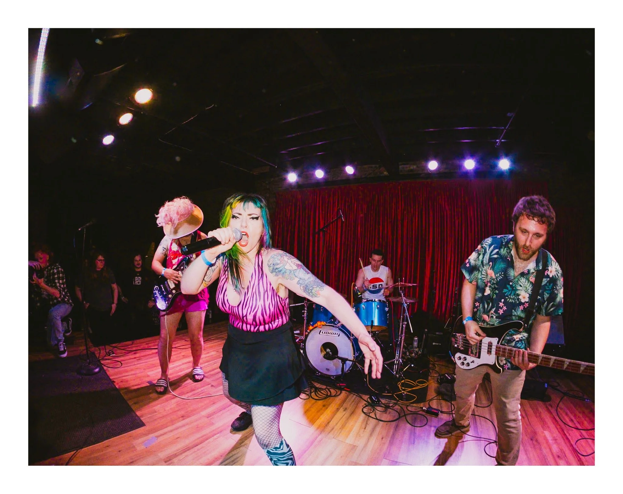 A band performs on stage with a female vocalist singing into a microphone, wearing tattoos and striped clothing, with band members playing guitars and drums in the background, illuminated by stage lights.