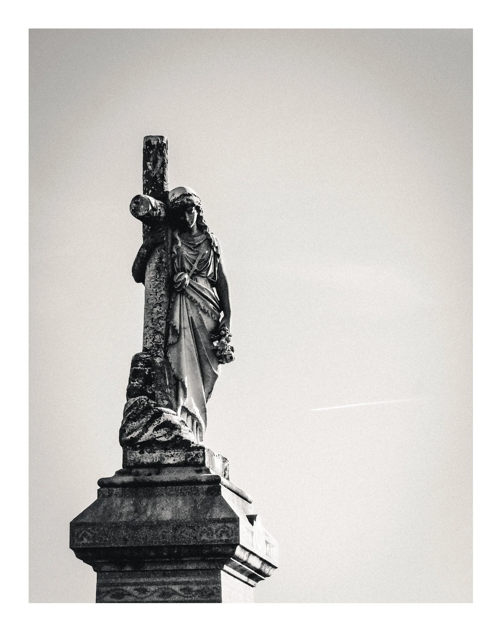 A black-and-white photograph of a weathered statue of a woman carrying a cross, standing on a stone pedestal against a clear sky.