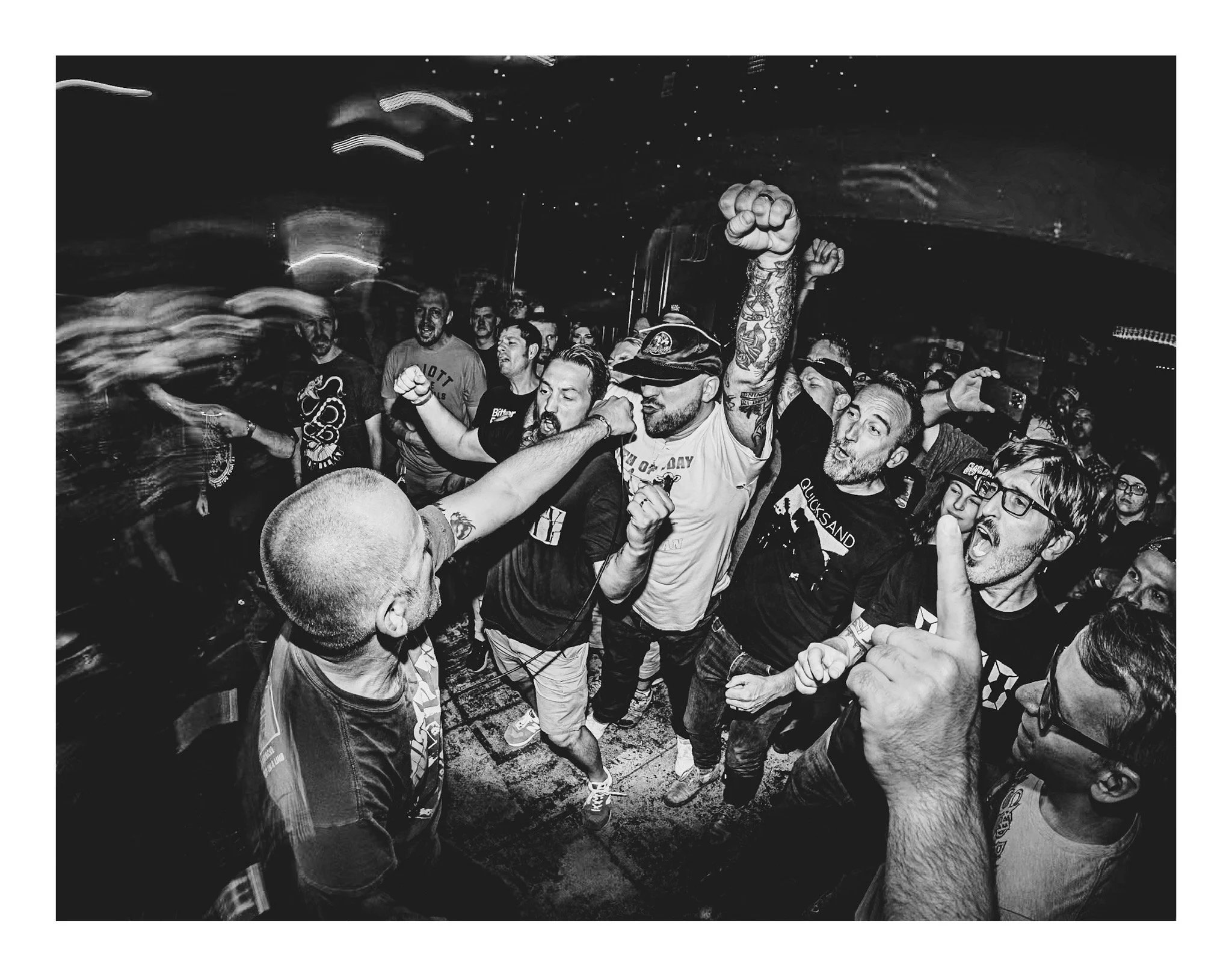 A group of people at a party or concert, some raising their fists in the air, with lively expressions and casual clothing, in a dimly lit environment with a dark ceiling.