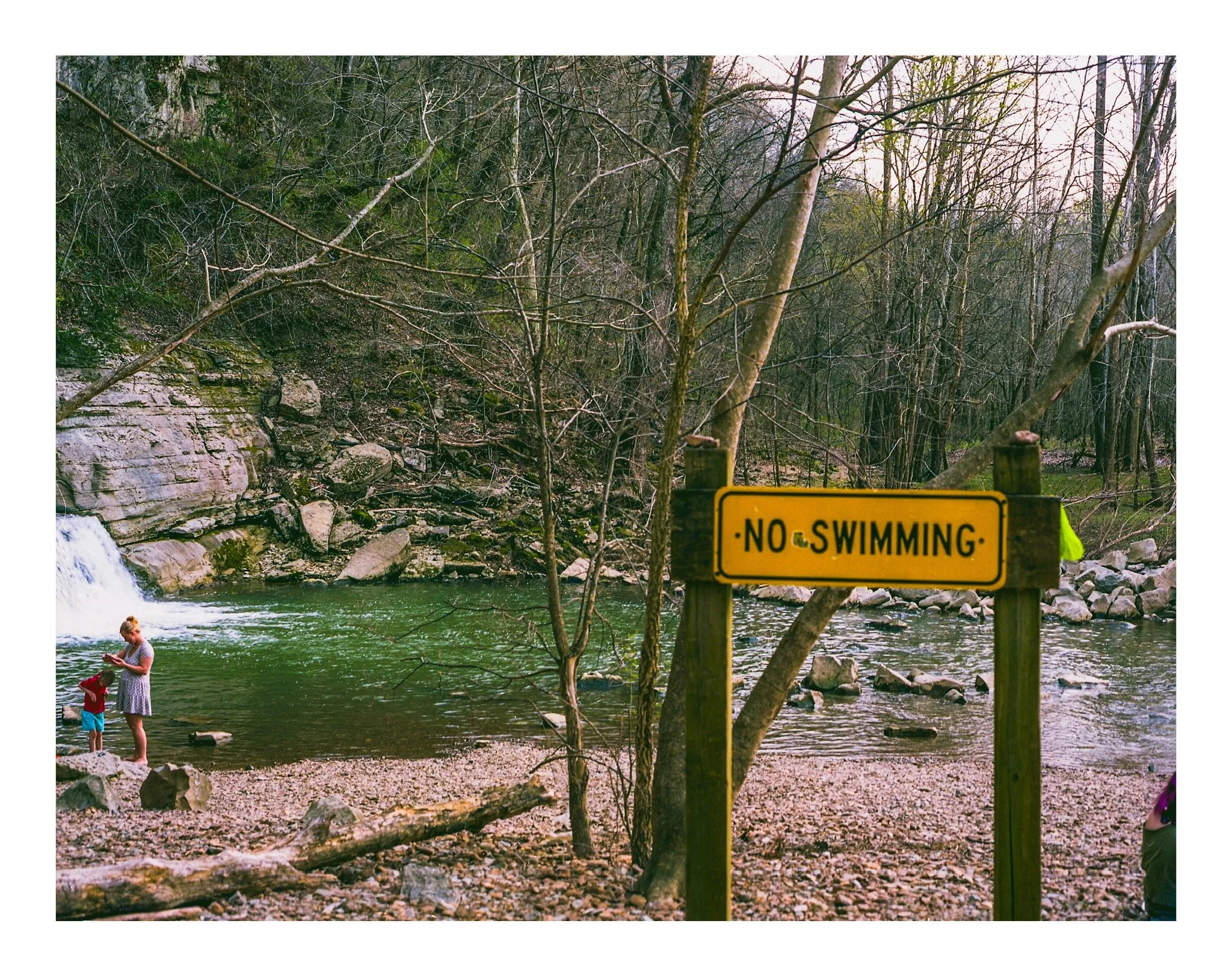 Children and a woman playing near a river with rocks, surrounded by trees and a yellow 'No Swimming' sign.