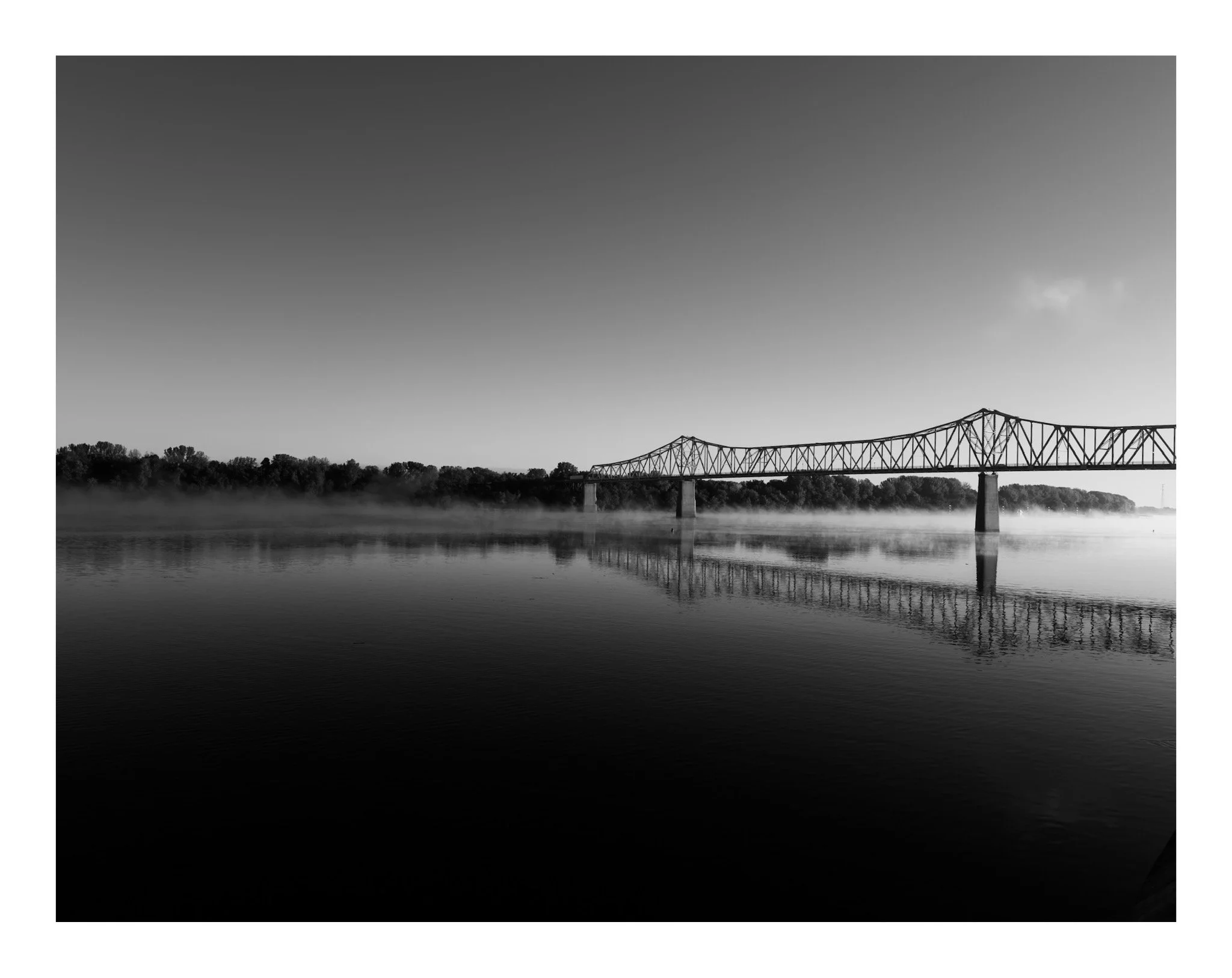 Black and white photograph of a bridge over a river with fog rising from the water, trees along the riverbank, and a clear sky with a few clouds.