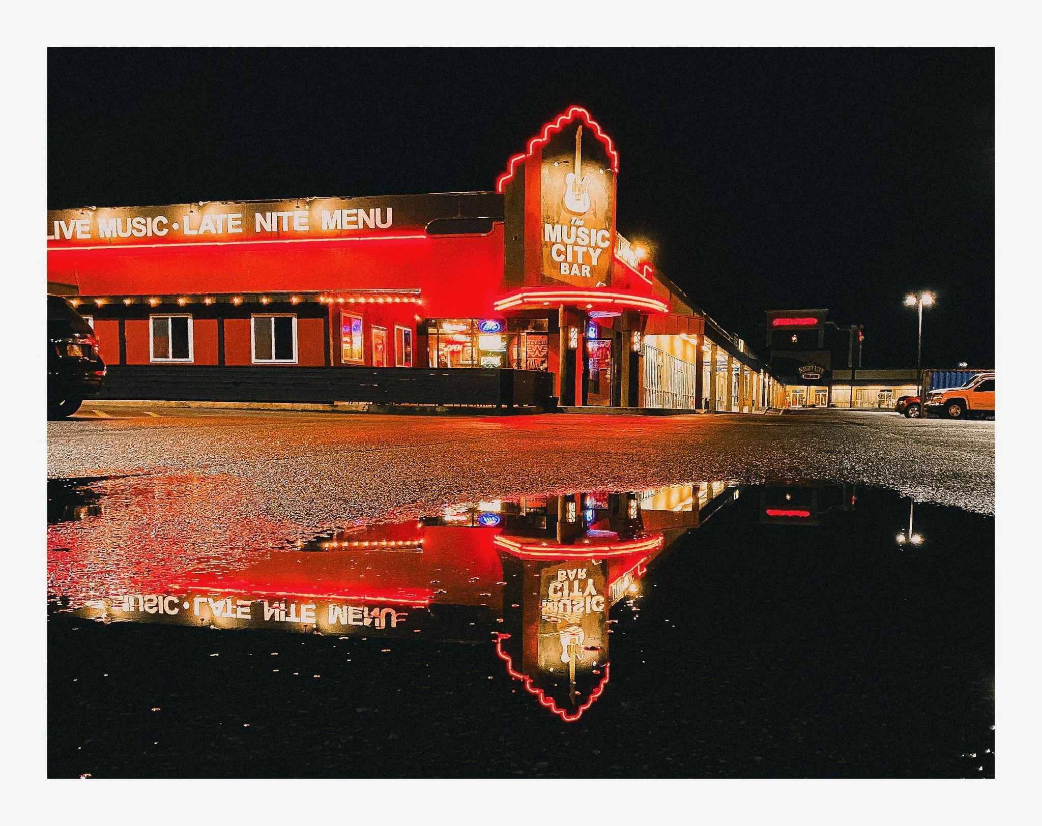 Nighttime view of a neon-lit music bar with signs advertising live music, a late night menu, and a guitar logo. Reflection of the sign is visible on a puddle in the parking lot.