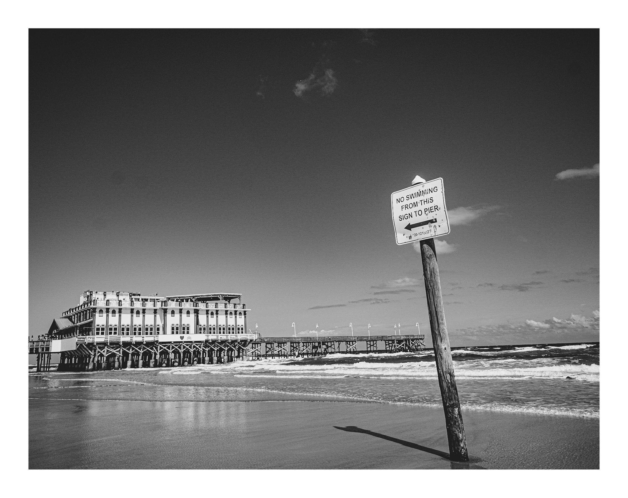 A black and white photo of a pier extending over the ocean with a building at the end. A sign on a wooden post in the foreground reads, 'No swimming from this sign to pier,' with an arrow pointing left.