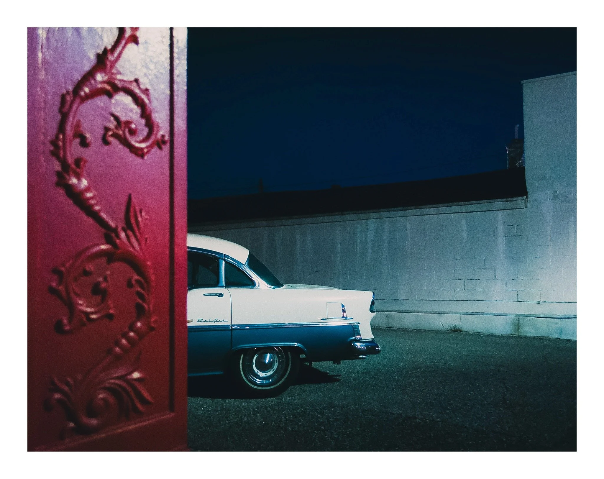 A vintage white car parked against a brick wall at night, with a red decorative metalwork piece partially visible on the left side of the image.