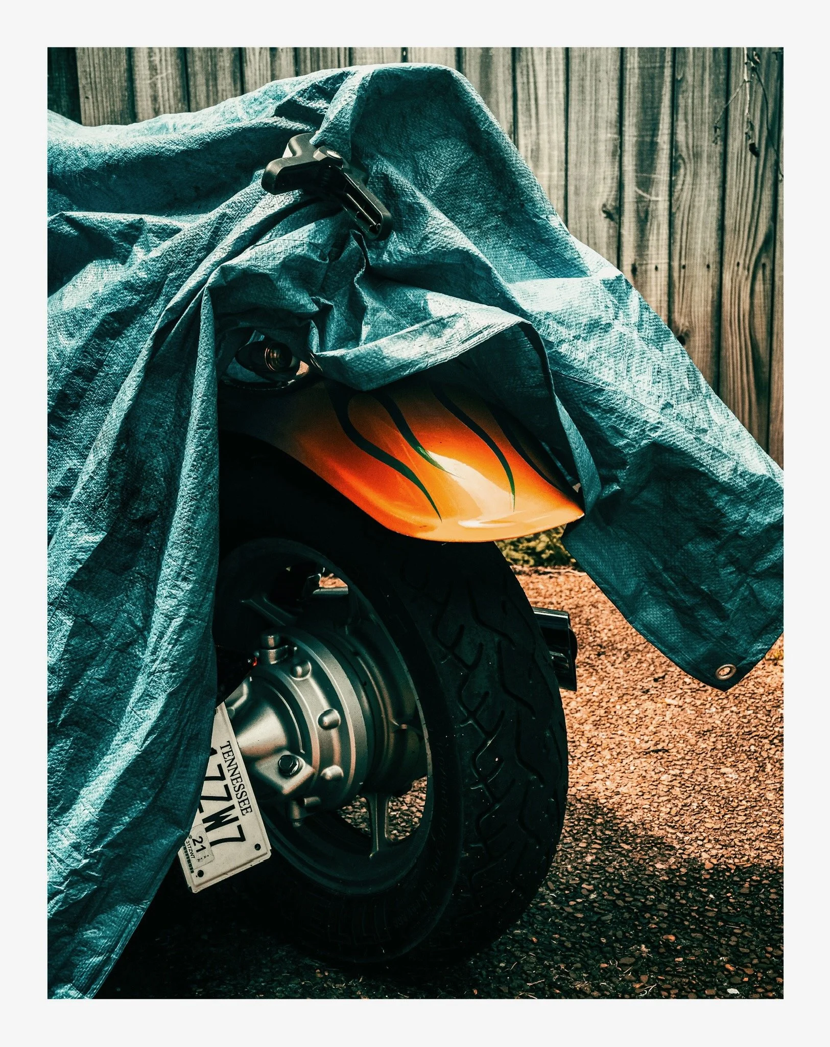 Close-up of a motorcycle tire and brake with a blue tarp partially covering it, set against a wooden fence background.