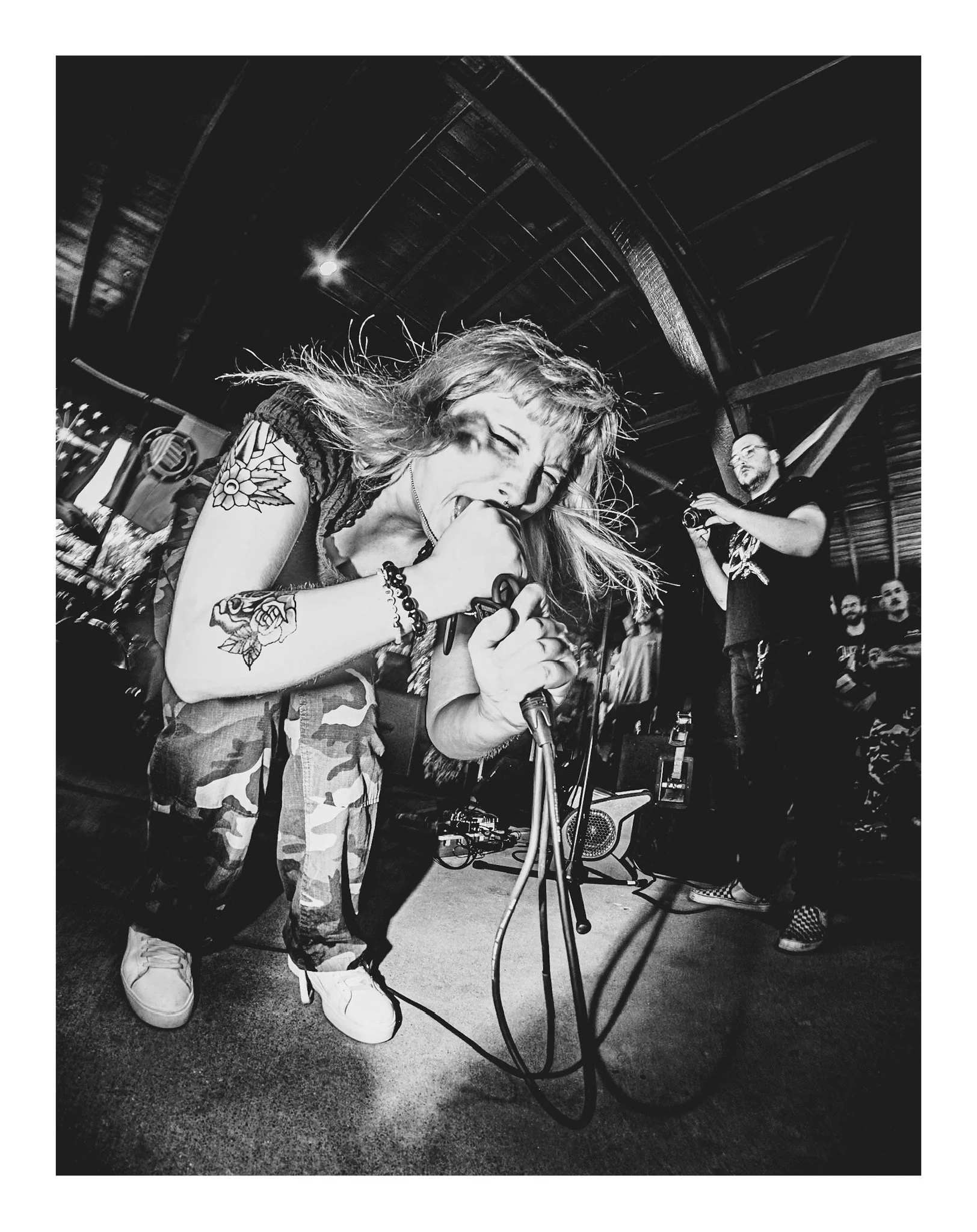 A woman with tattoos on her arm singing into a microphone with a man in the background holding a camera. The scene appears to be in a dimly lit venue with a curved ceiling.