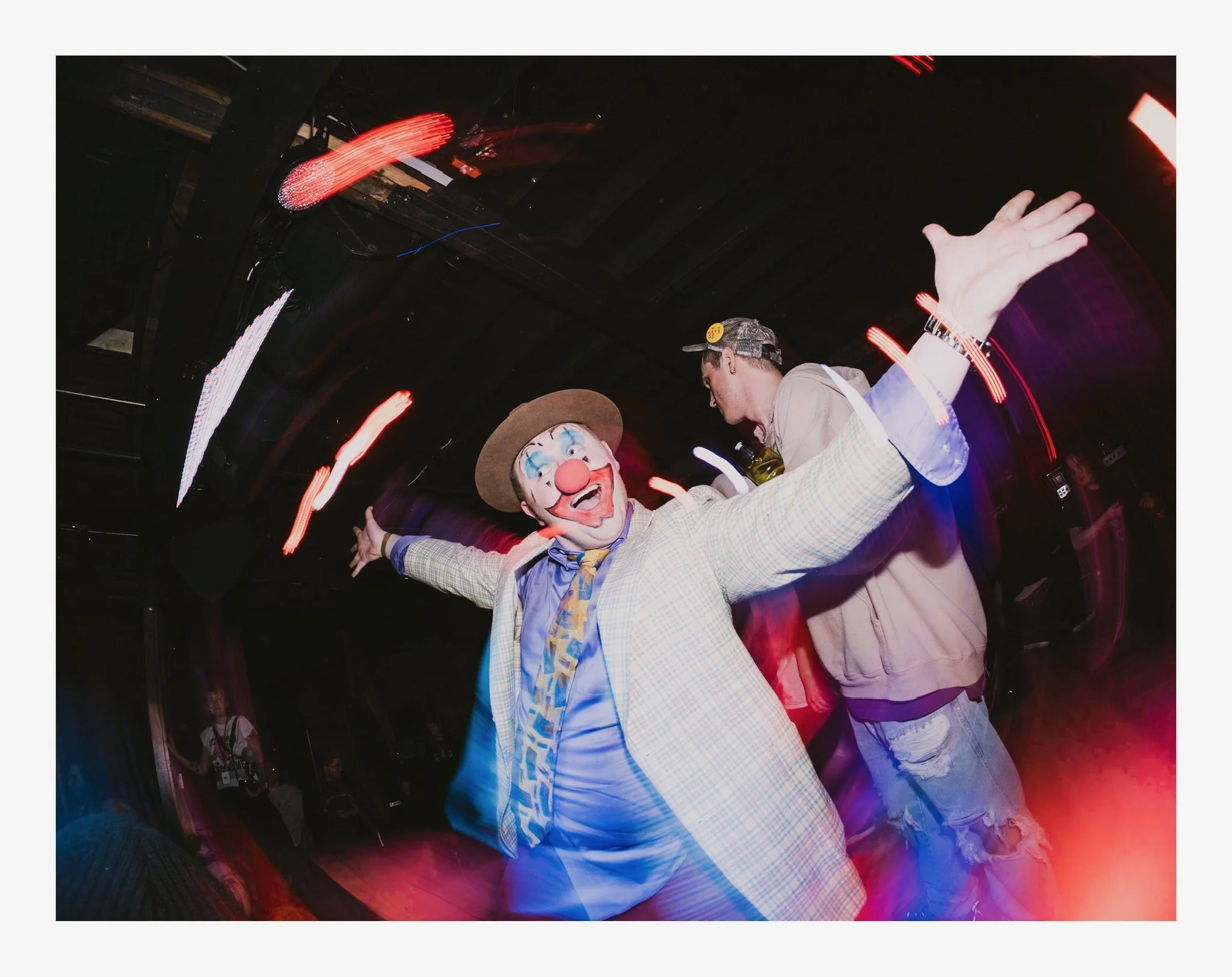 A person in a clown costume with a painted face, large red nose, and a plaid suit is dancing with arms outstretched at a party or celebration, with colorful lights and other people in the background.
