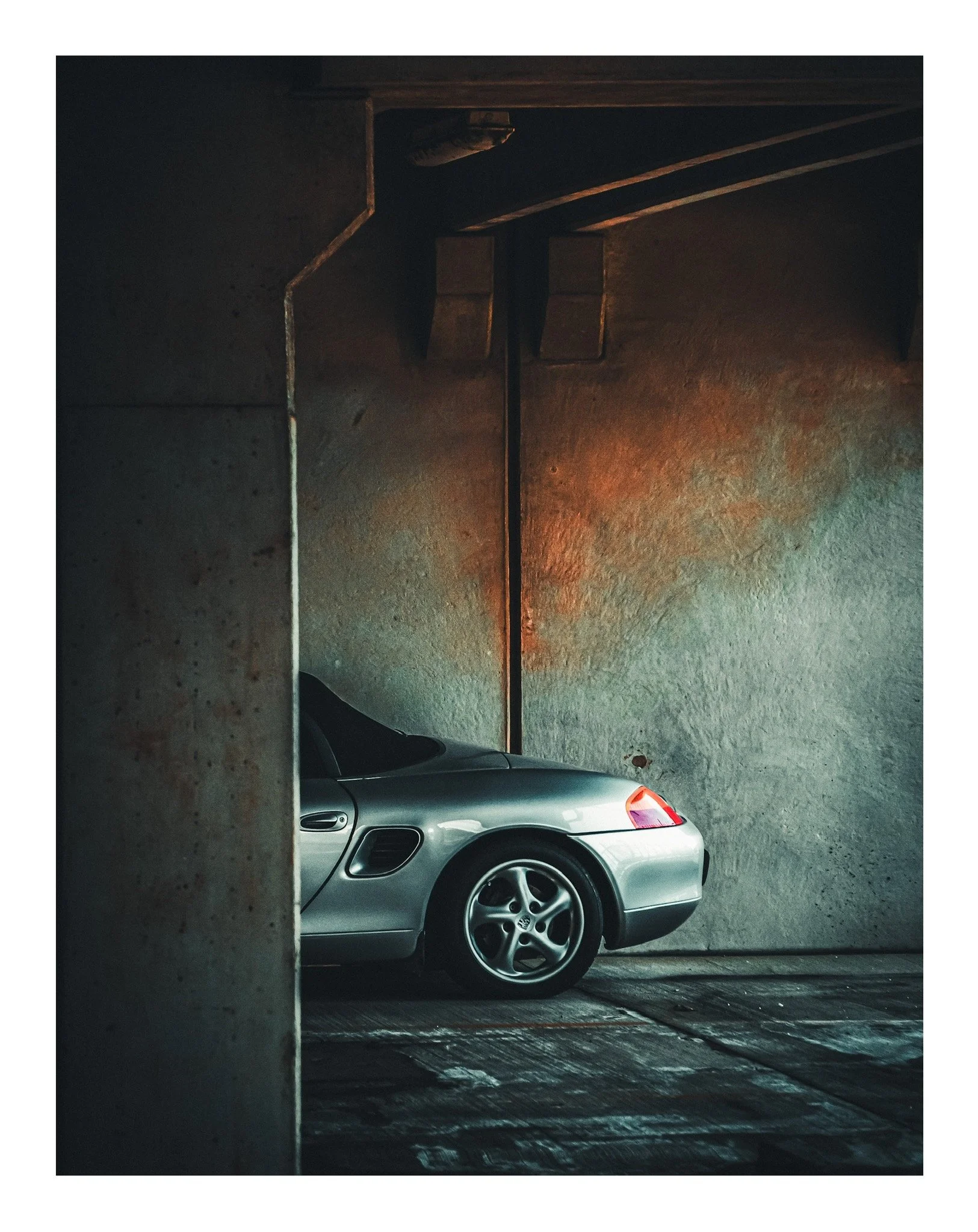 Silver sports car partially visible behind a concrete pillar in a dimly lit parking garage with textured concrete walls.