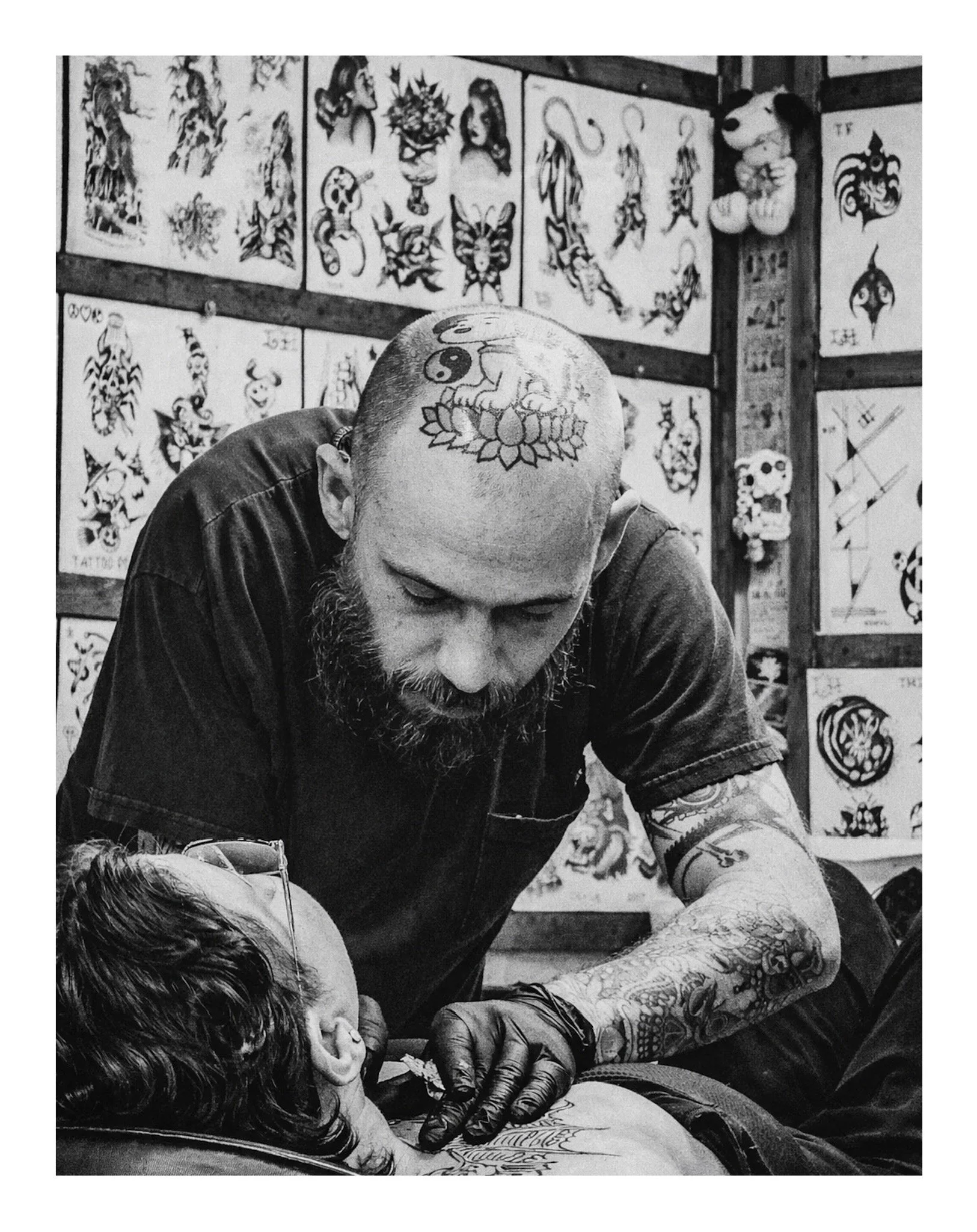 A man with a shaved head and tattoos on his arms, including a yin-yang symbol on his forehead, is tattooing a person's arm while another person is lying with their eyes closed in front of him. The background shows tattoo designs on display.