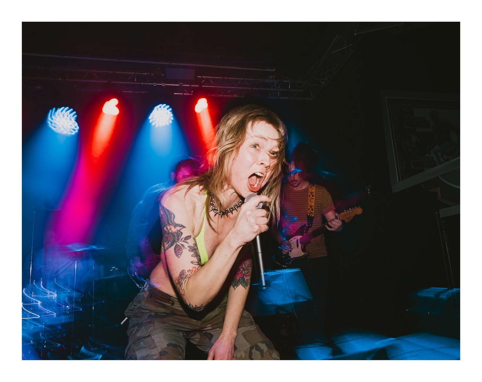 A woman singing into a microphone on stage with colorful lights behind her, a band member playing guitar in the background.