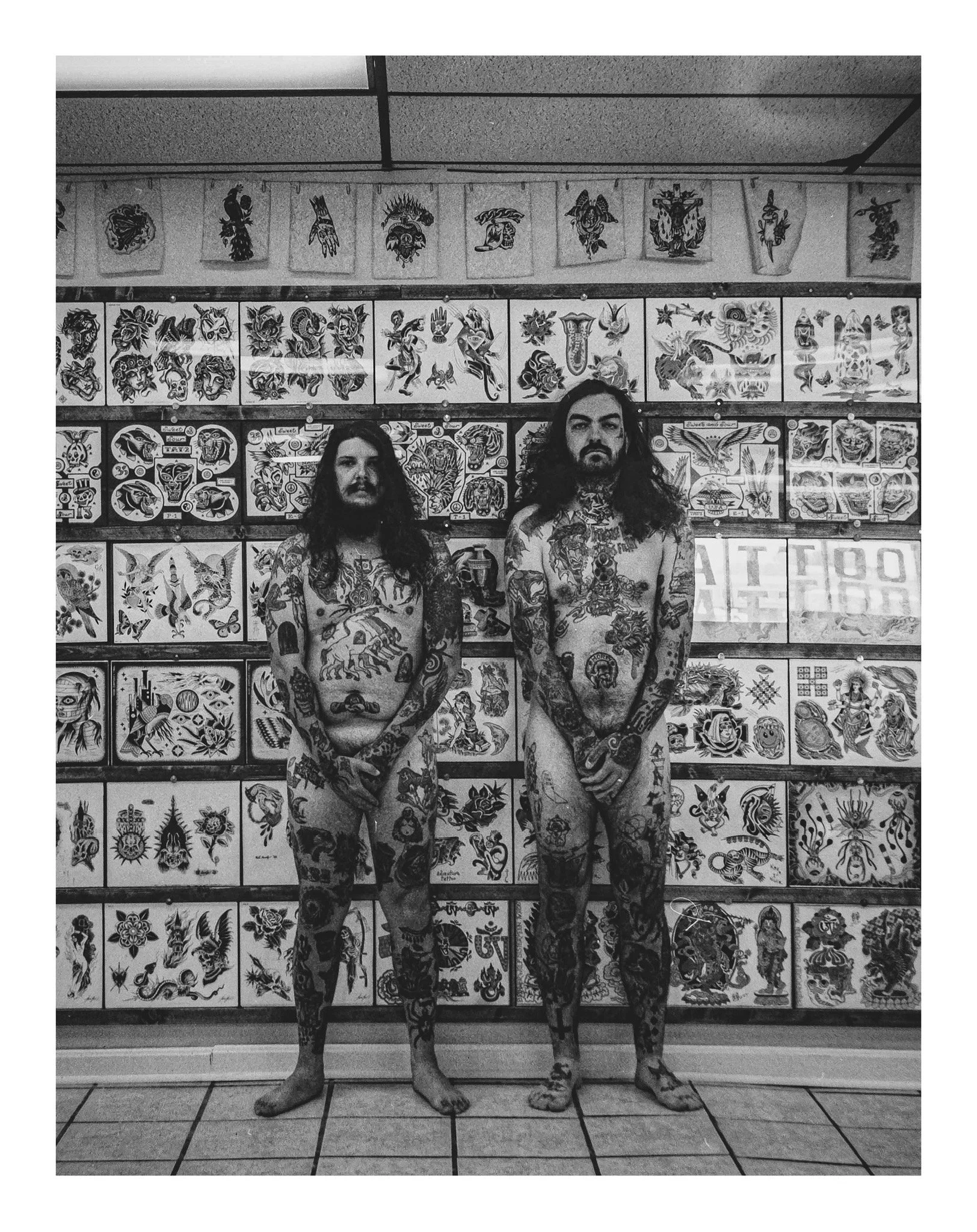 Two tattooed men with long hair standing shirtless in front of a wall covered with tattoo flash art, with tiled floor and ceiling tiles visible.