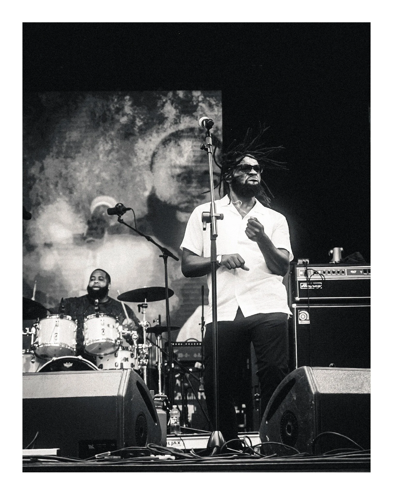 A black and white photo of a musician with dreadlocks and sunglasses singing on stage. A drummer is seated behind him, and stage equipment including speakers and an amplifier are visible.